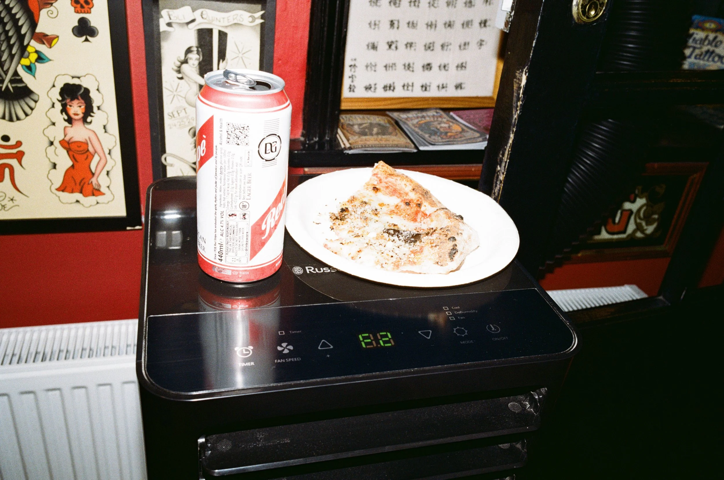 A slice of pizza on a white plate and a can of soda placed on a black induction cooker in a restaurant setting with various posters and menu items on the wall in the background.