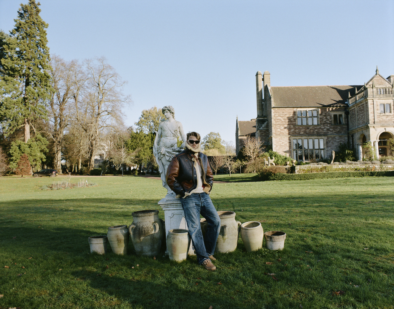 A man wearing sunglasses, a leather jacket, and jeans standing outdoors on a grass lawn next to a classical white marble statue of a woman, with old pottery pots arranged at his feet, in front of a large historic house under a clear blue sky.