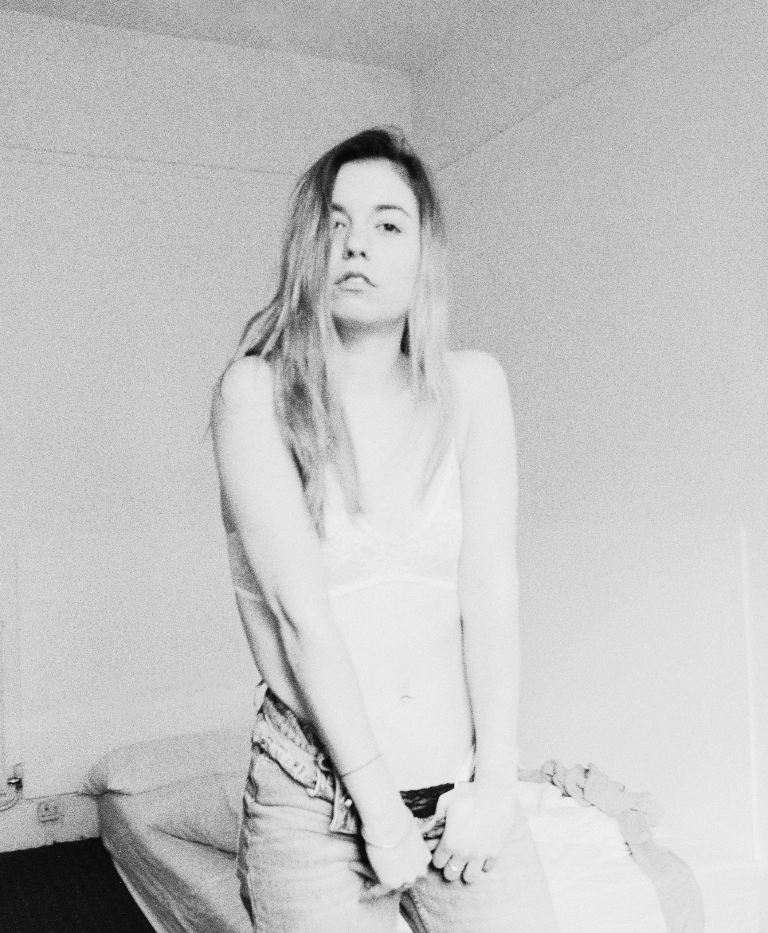 Black and white photo of a young woman with long hair, wearing a tank top and jeans, standing in a bedroom with a bed and some clothes in the background.
