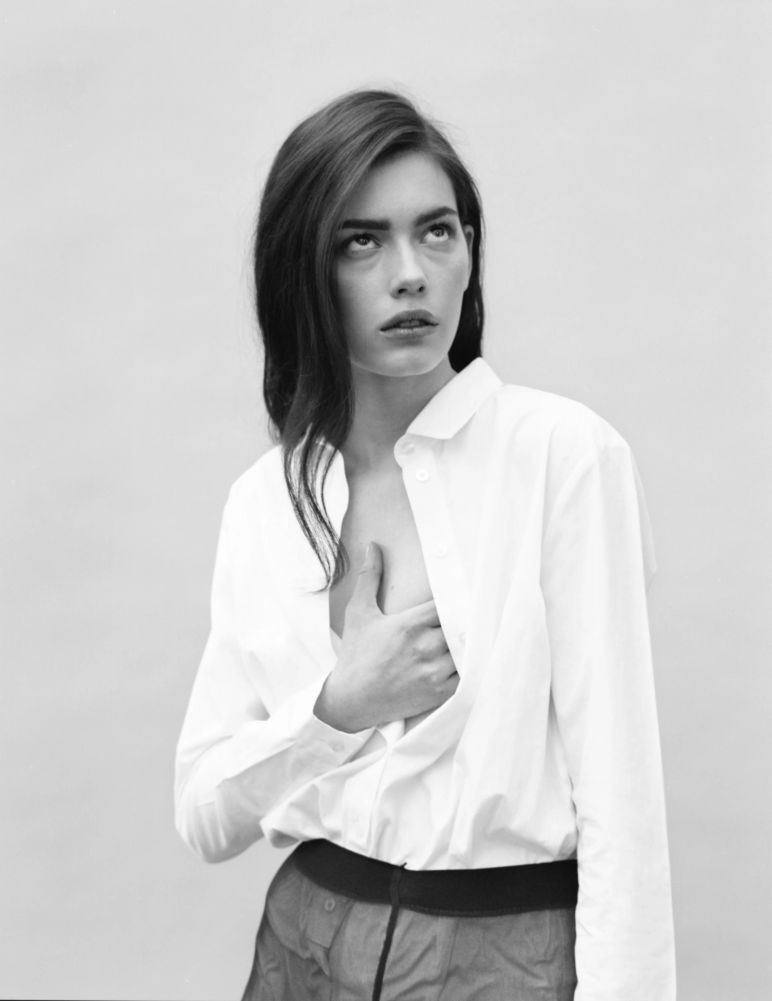 Black and white photo of a woman with shoulder-length dark hair, wearing a white button-up shirt, holding her chest, looking slightly upward and to the left.
