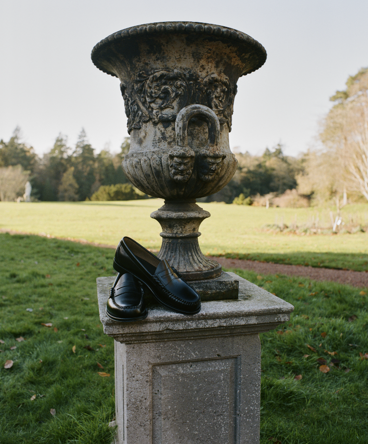 A pair of black dress shoes placed on a stone pedestal beneath an ornate stone urn in a park with green grass and trees in the background.