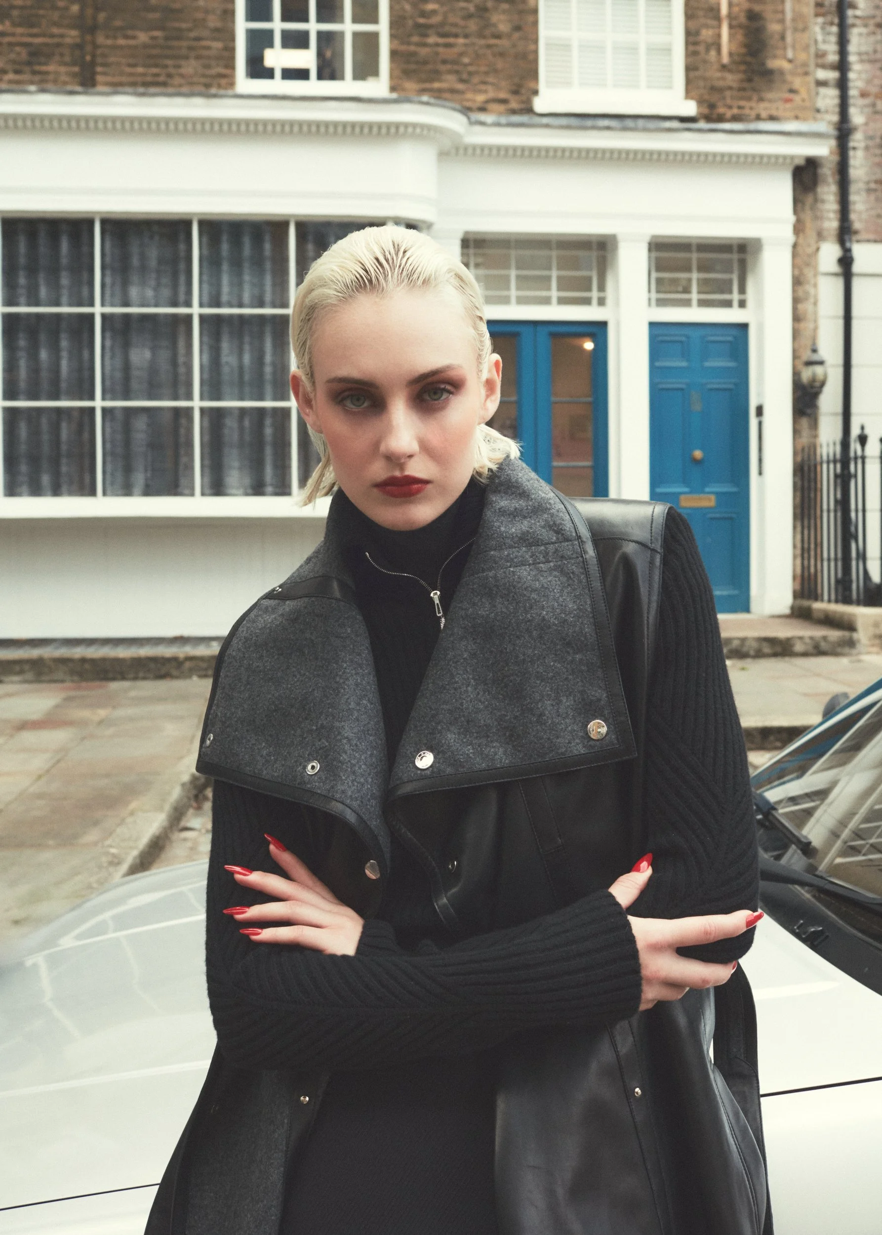 A woman with platinum blonde hair and red lipstick standing in front of a white car and a house with a bright blue door.