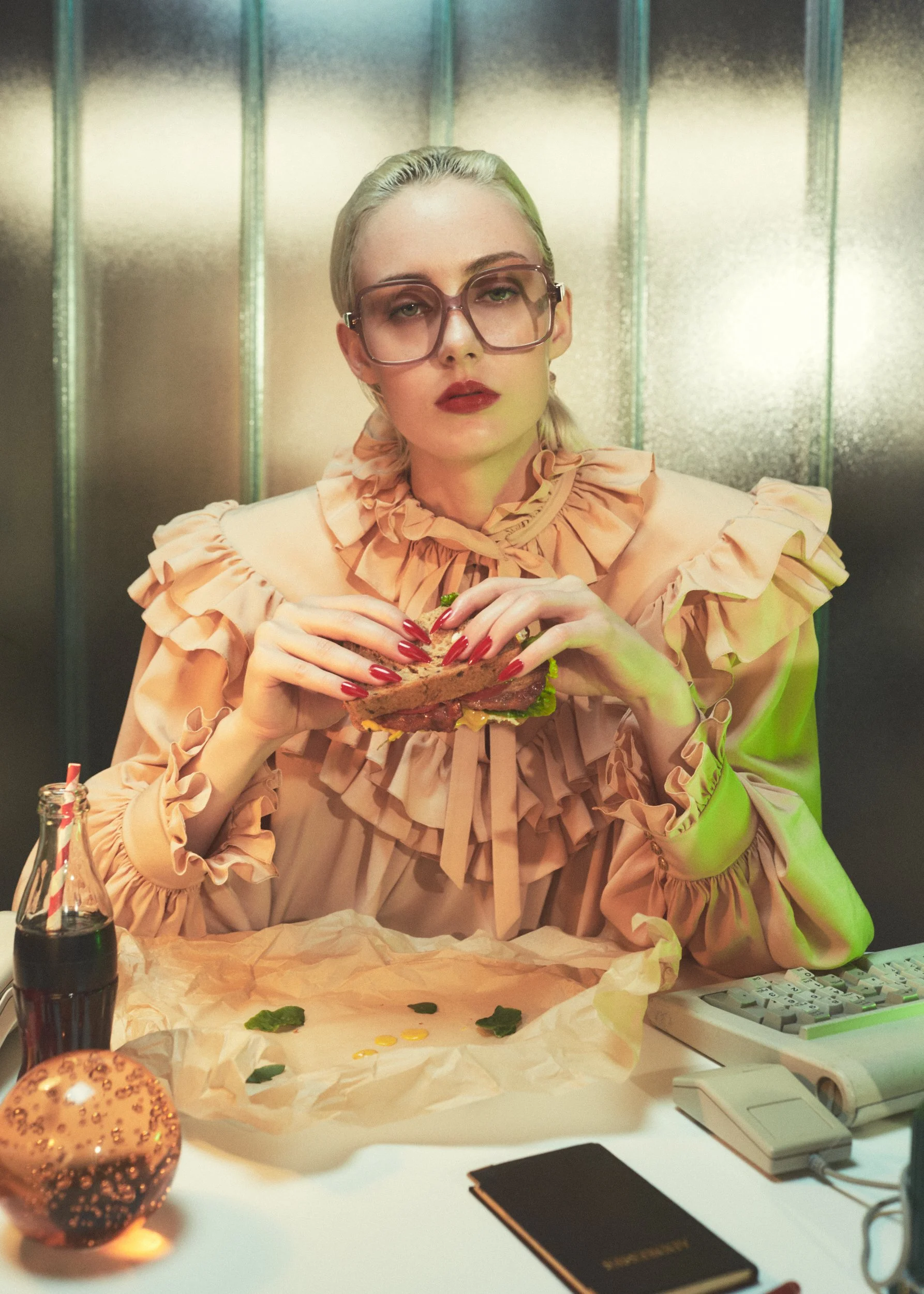 A woman with blonde hair, glasses, and red lipstick sitting at a desk, holding a sandwich with lettuce and meat. The desk has a soda bottle, a vintage keyboard, a notepad, and a decorative lamp. The woman is wearing a ruffled peach-colored blouse wit