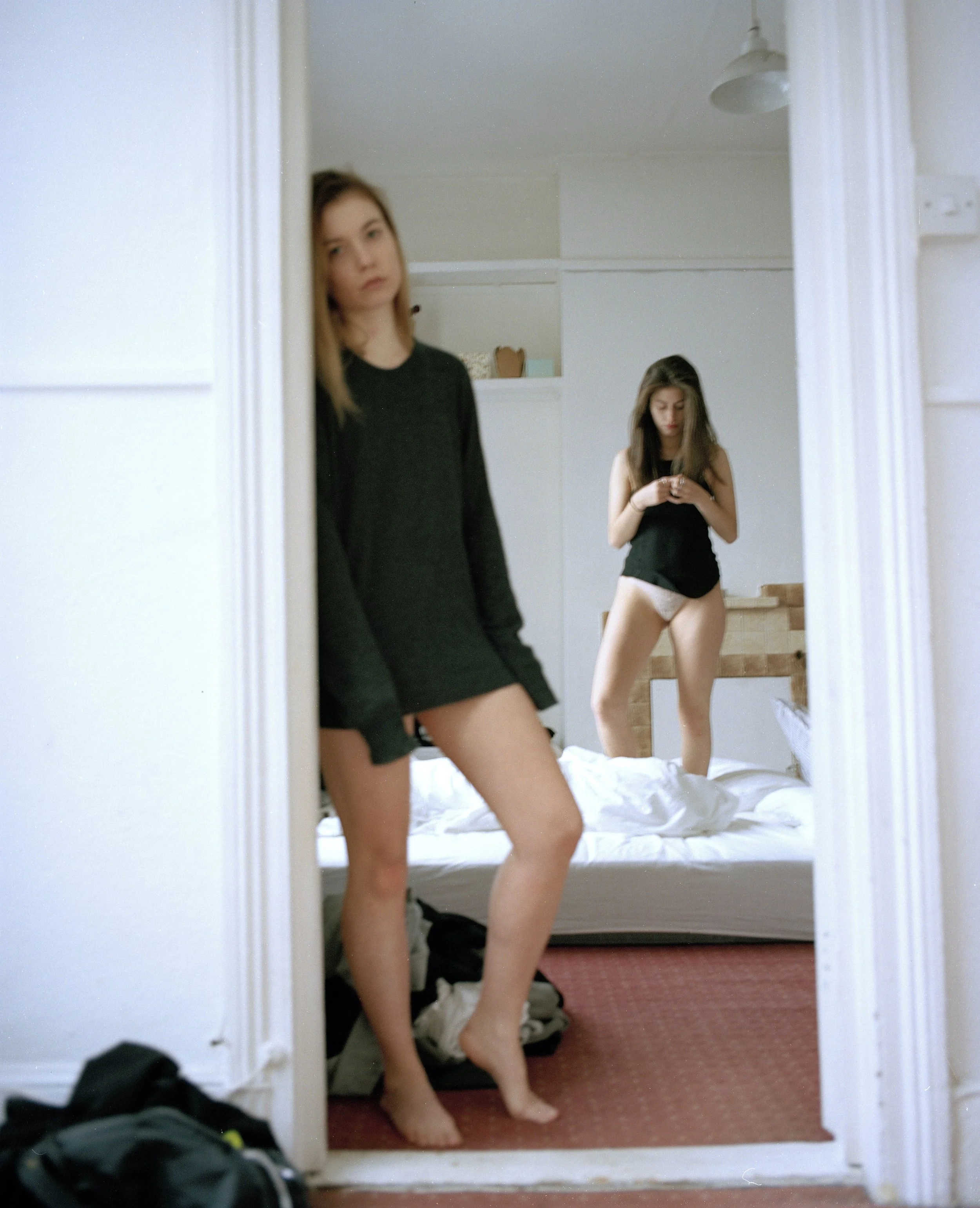 Two young women standing in front of a mirror in a bedroom. One is looking at the camera, and the other is looking at her phone. The woman in the foreground is wearing a black long-sleeve shirt and white underwear, while the woman in the background i