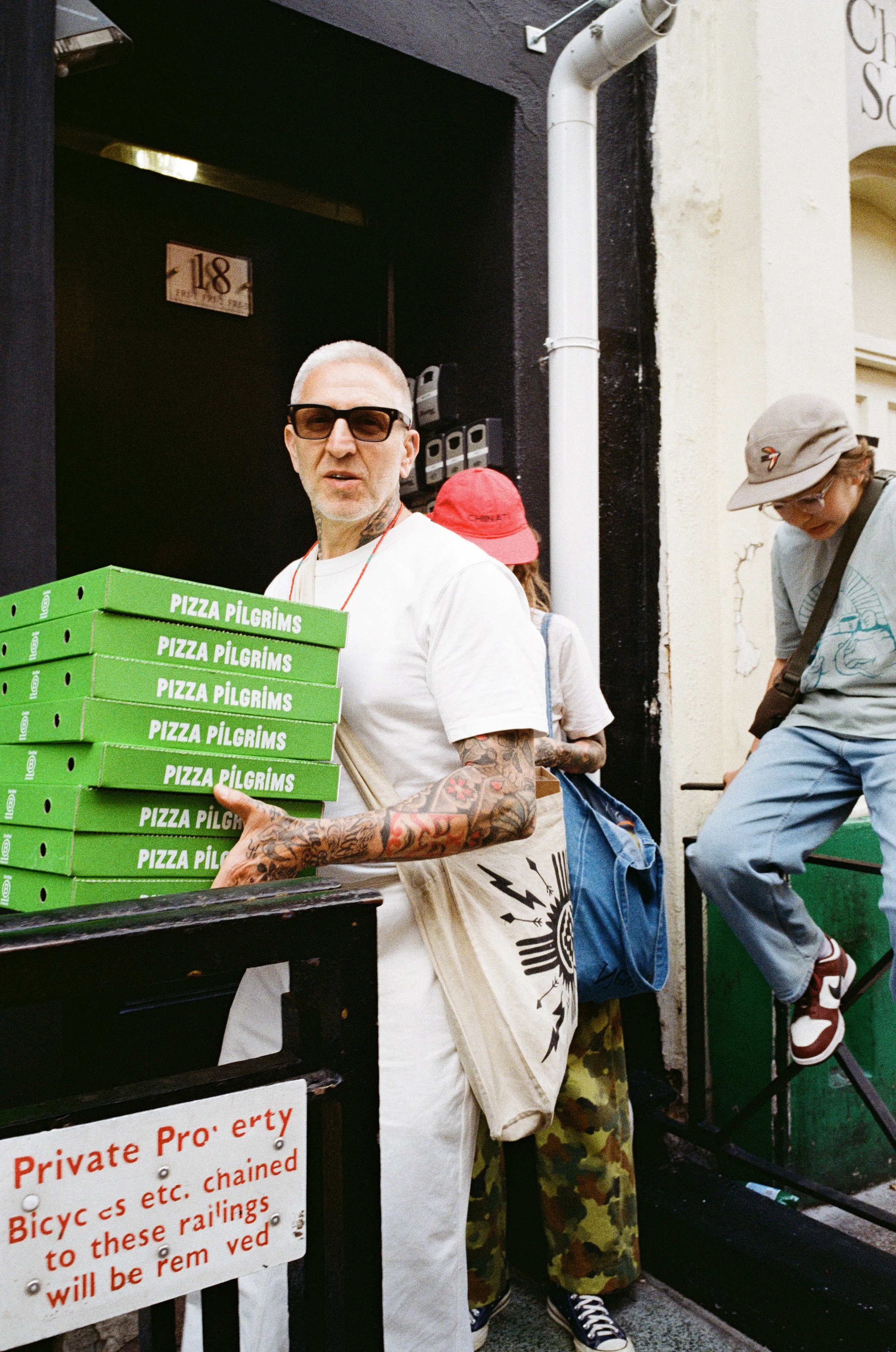 A man with tattoos and sunglasses holding multiple pizza boxes labeled "Pizza Pilgrims" outside a building, with a partially visible person wearing camouflage pants and another person on stairs in the background.