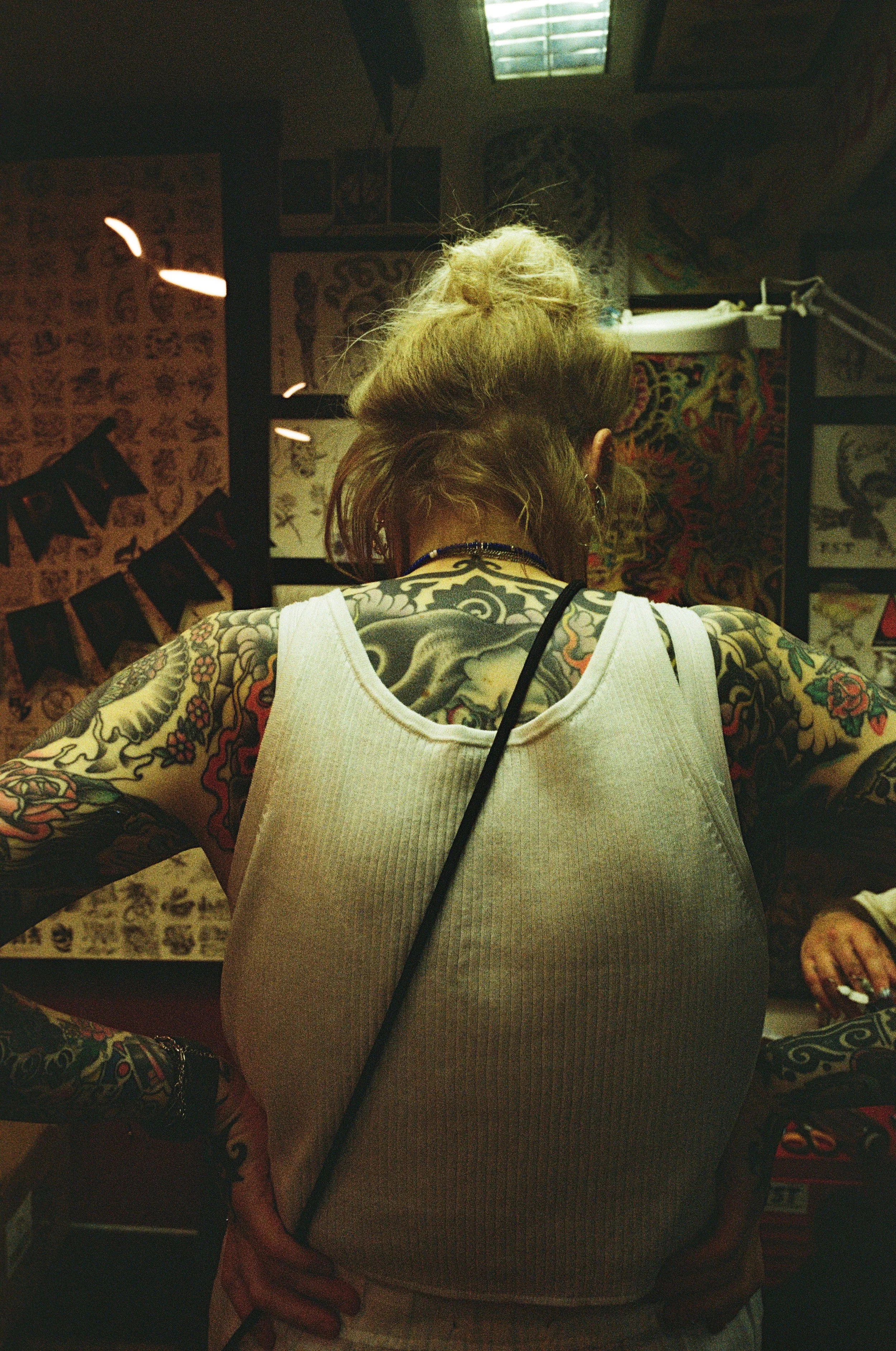 Back of a woman with patterned tattoos, wearing a white sleeveless top, standing in a room decorated with tattoos and artwork.