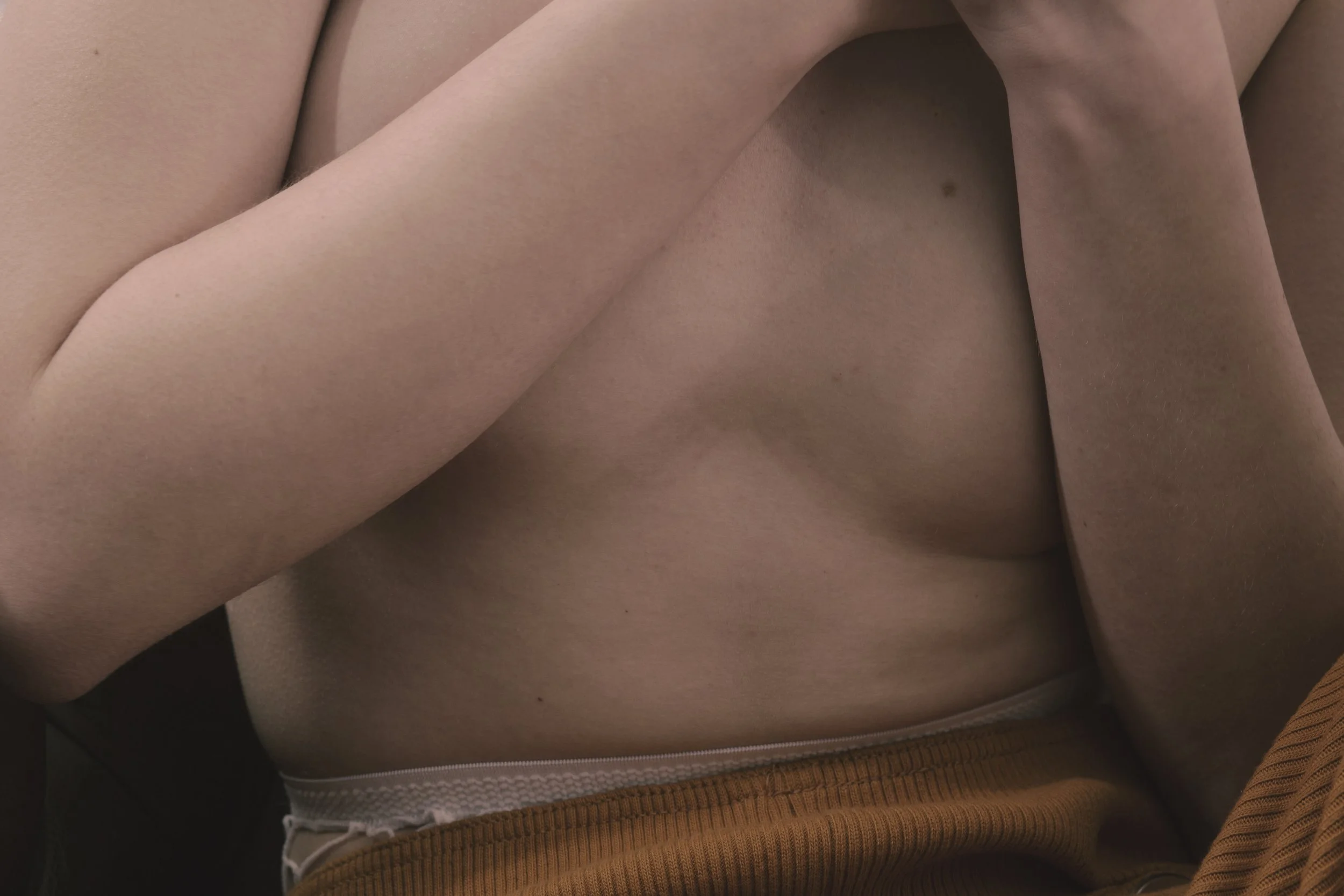 Close-up of a person’s bare chest with one arm crossing the body, and a partial view of orange clothing at the bottom.