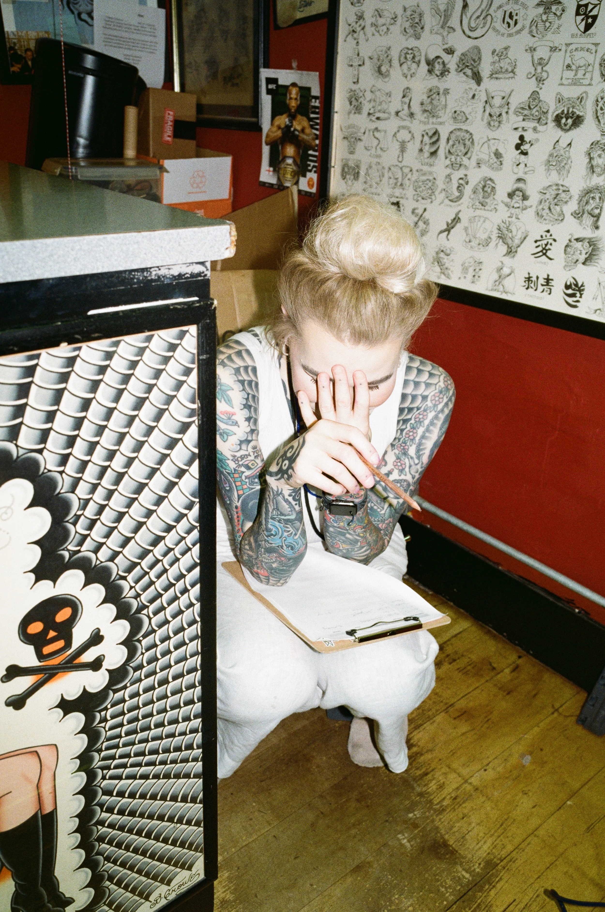 A woman with colorful tattoos, blonde hair styled in a vintage updo, sits on a small stool in a tattoo shop. She holds a pencil and a clipboard, appearing to be in deep thought or sketching. The shop has tattoo flash art on the wall, and a menu or li