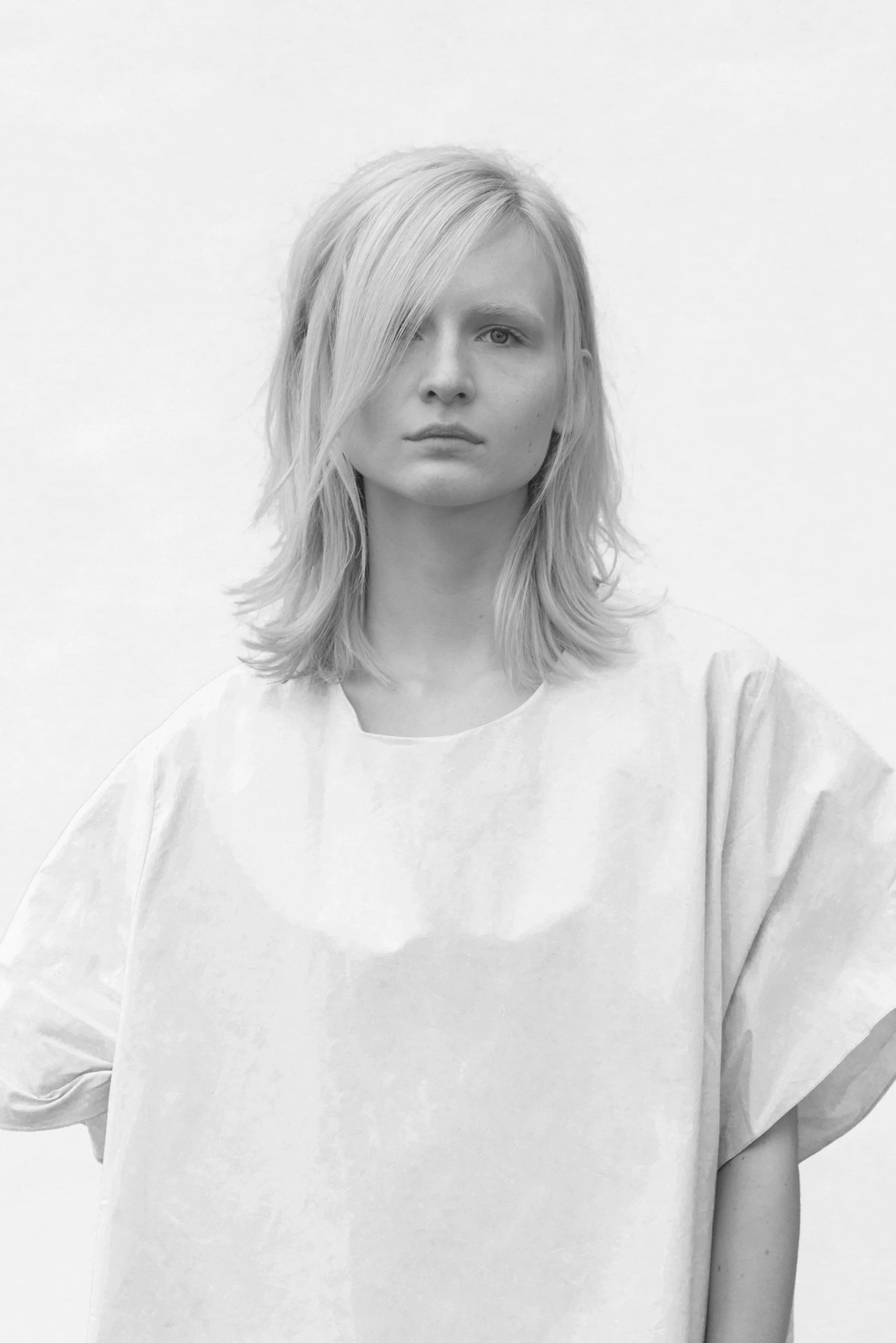 Black and white photo of a woman with shoulder-length blond hair, wearing a loose light-colored shirt, looking directly at the camera with a serious expression.