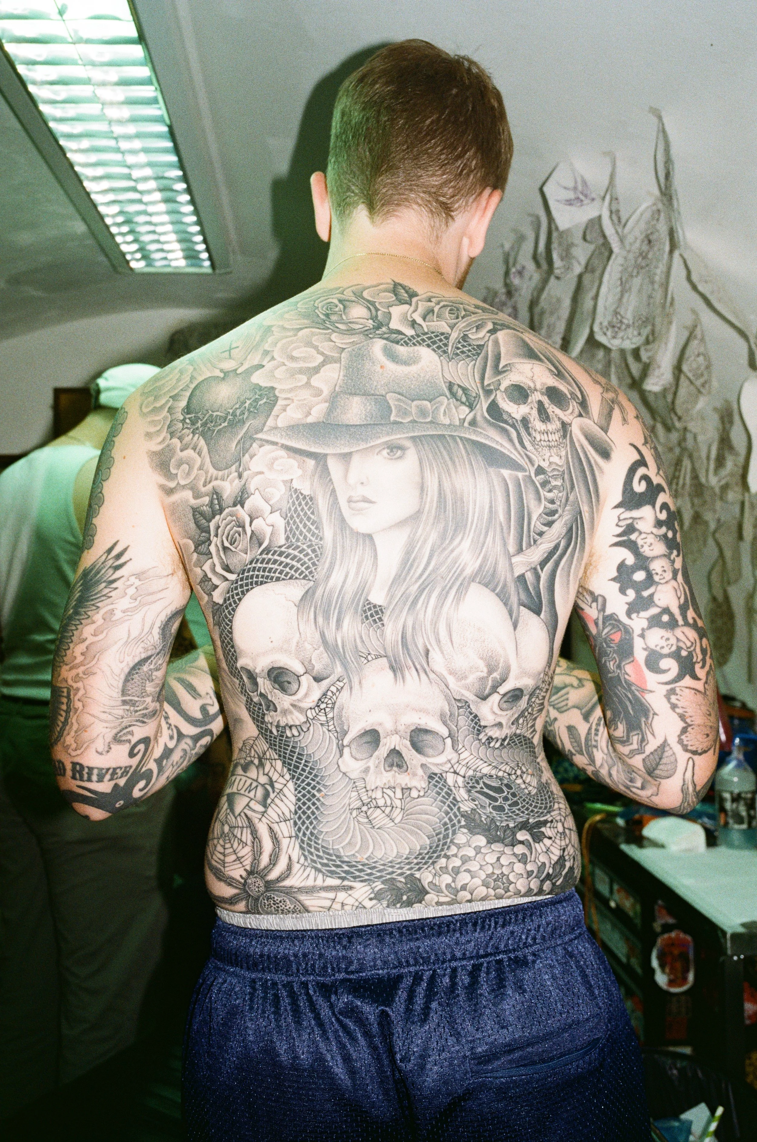 A man with a full back tattoo of skulls, a snake, roses, clouds, and a woman wearing a witch's hat with long hair.