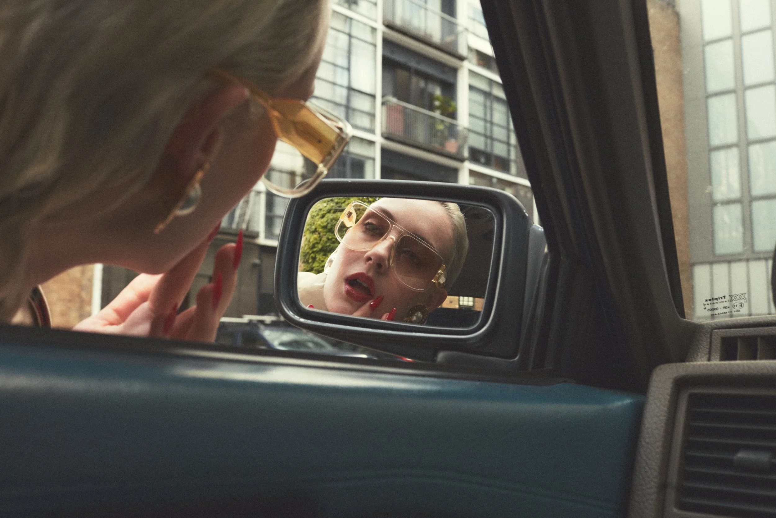 A woman with blonde hair and gold sunglasses looks into a car's side mirror. Her reflection is visible in the mirror, showing her face with makeup and red lipstick as she touches her face with her fingers. The background shows an urban apartment building with balconies.
