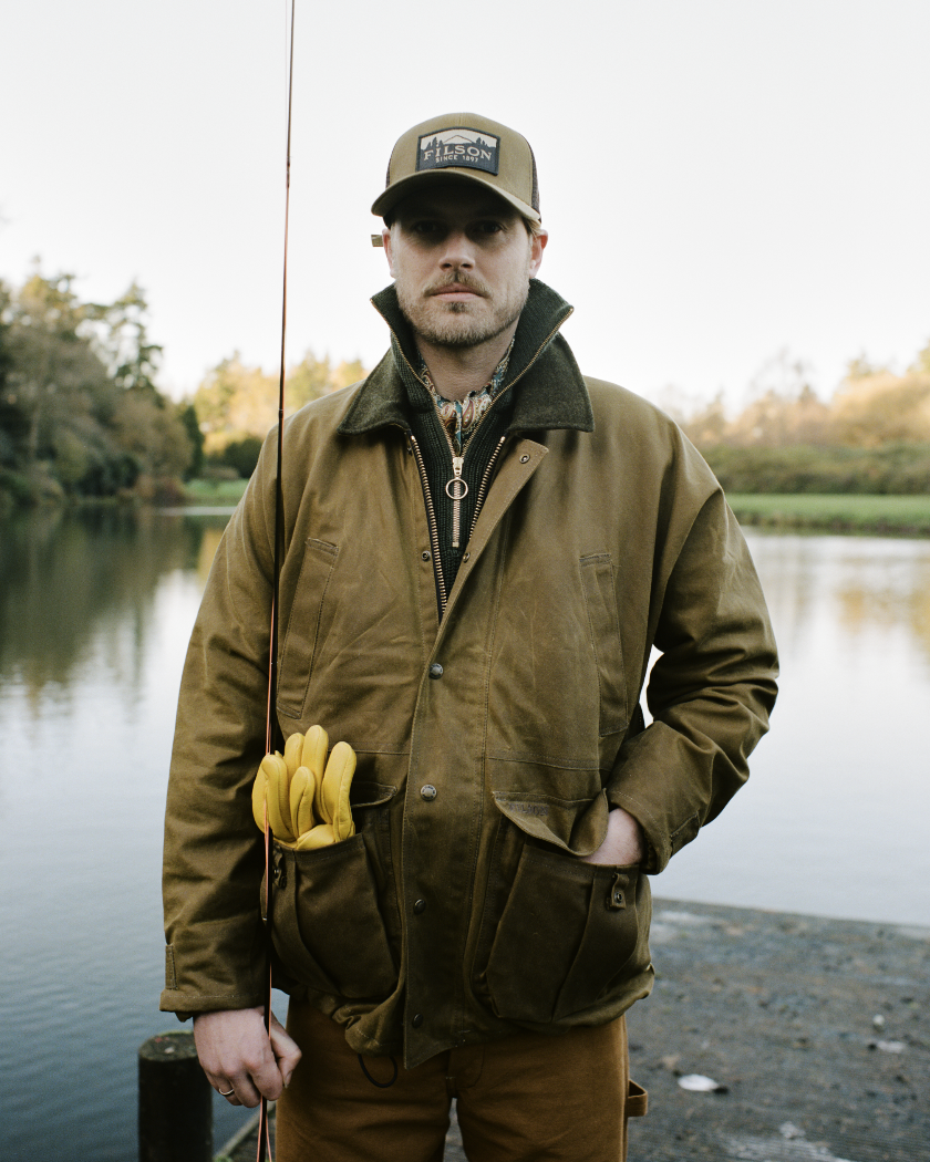 Man standing outdoors by a river holding a fishing rod with yellow gloves attached to his jacket pocket, wearing a brown jacket and a cap, with trees in the background.