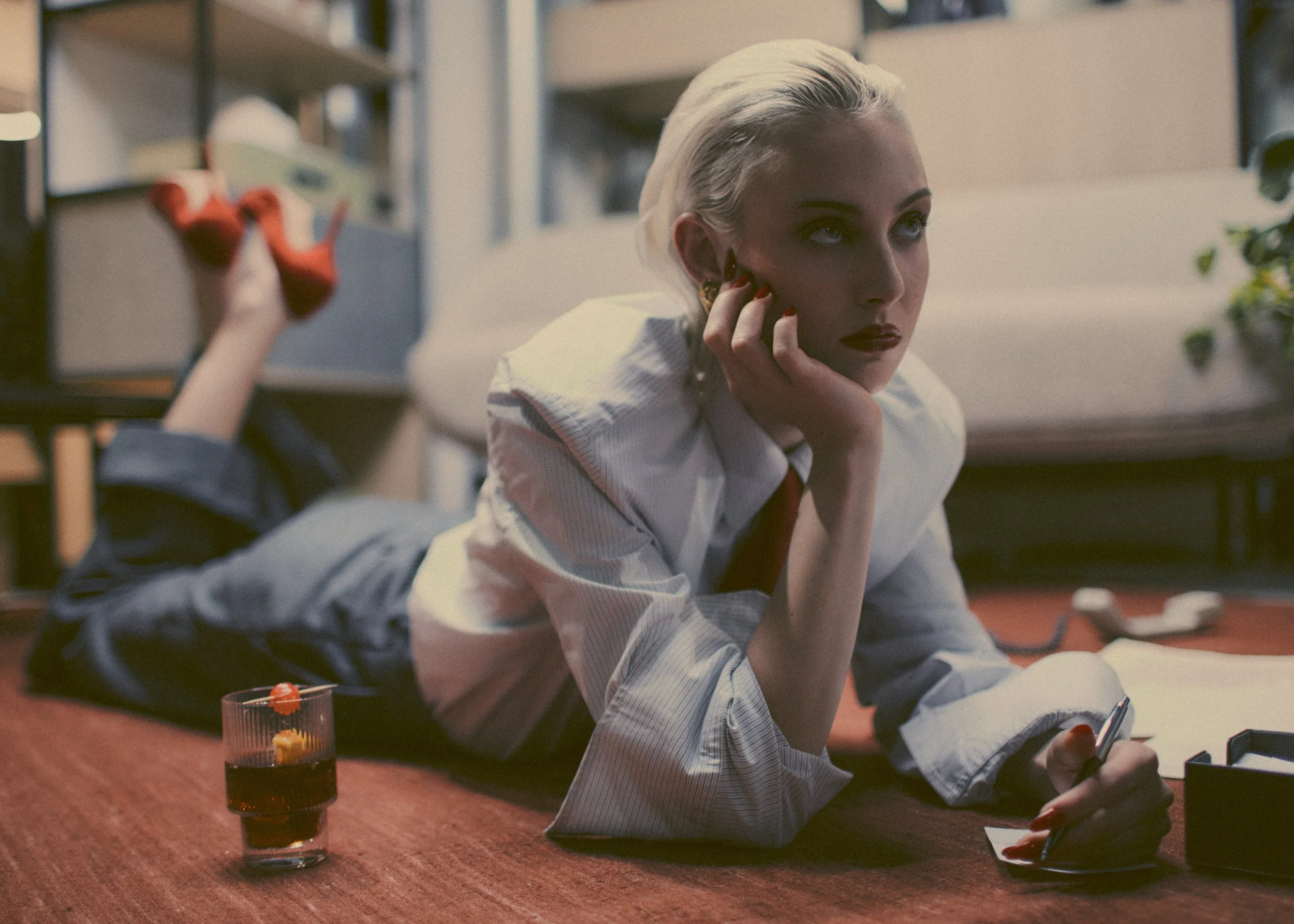 A woman with platinum blonde hair lying on her stomach on a red carpeted floor, resting her head on her hand, holding a pen in her other hand, with a glass of dark beverage and some papers nearby, in a room with shelves and plants.