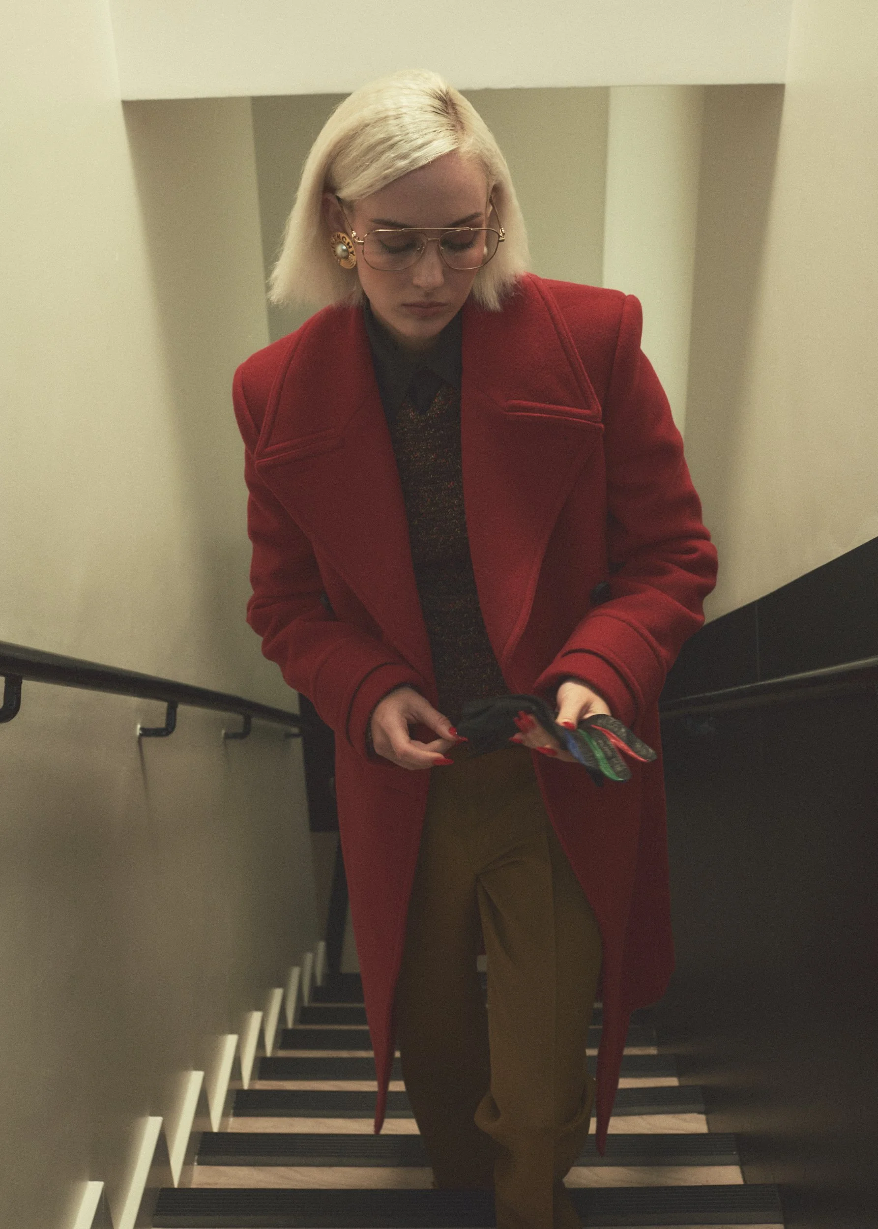 A woman with blonde hair, glasses, and earrings wearing a red coat walking down stairs, looking at her phone.