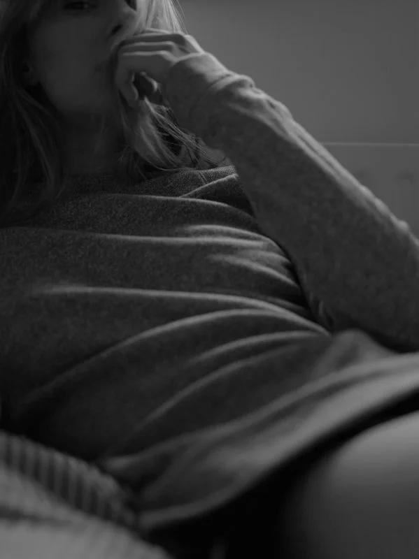A woman with long hair, wearing a long-sleeved shirt, is sitting and resting her hand near her face in a black-and-white photo.