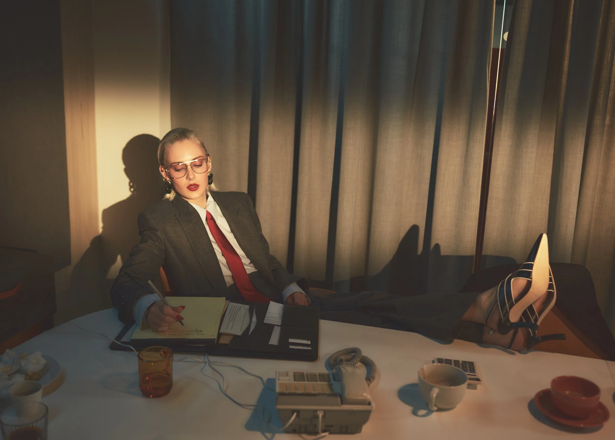 A woman in formal business attire, including a blazer, white shirt, and red tie, is sitting in an office with her legs stretched out on the desk. She is writing in a notebook, wearing glasses, and has her eyes closed. There are various office items o