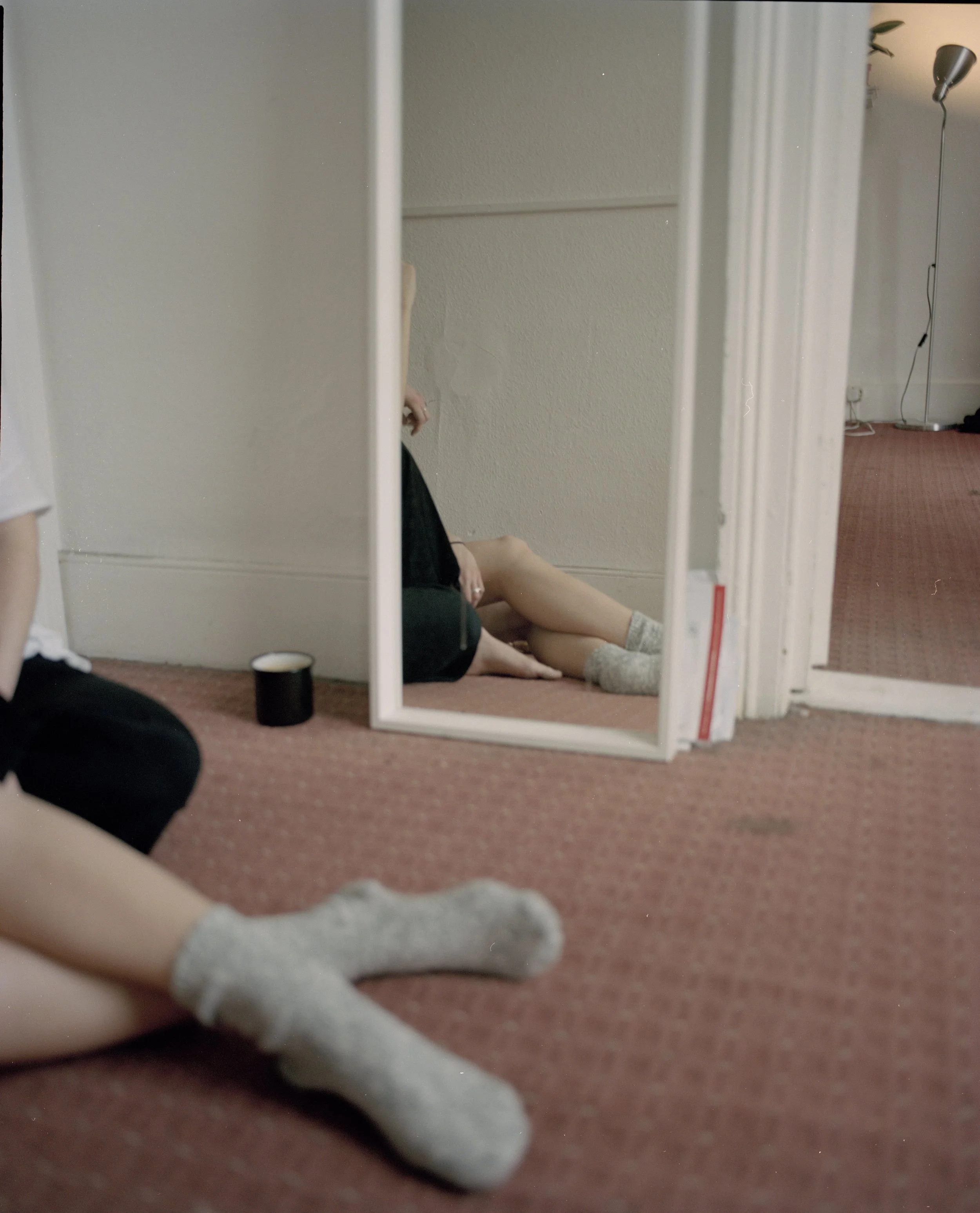 A person wearing socks, sitting on a red carpeted floor, taking a photo of themselves reflected in a full-length mirror leaning against the wall. The reflection shows their legs and part of their arm, with a cup placed nearby on the floor. The room h