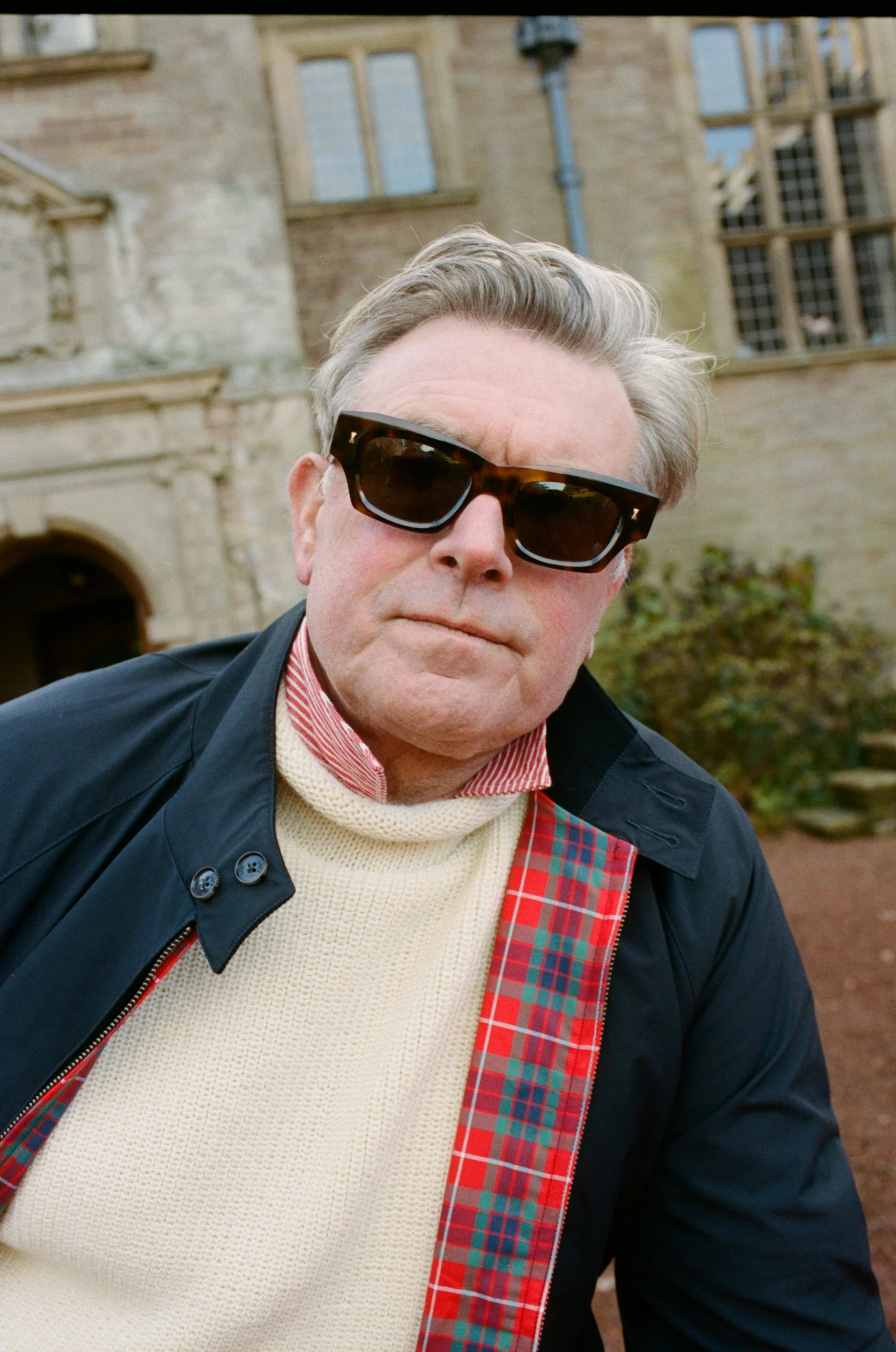 An older man wearing dark sunglasses, a cream sweater, and a dark jacket with red plaid lining stands outdoors in front of an old stone building with large windows.