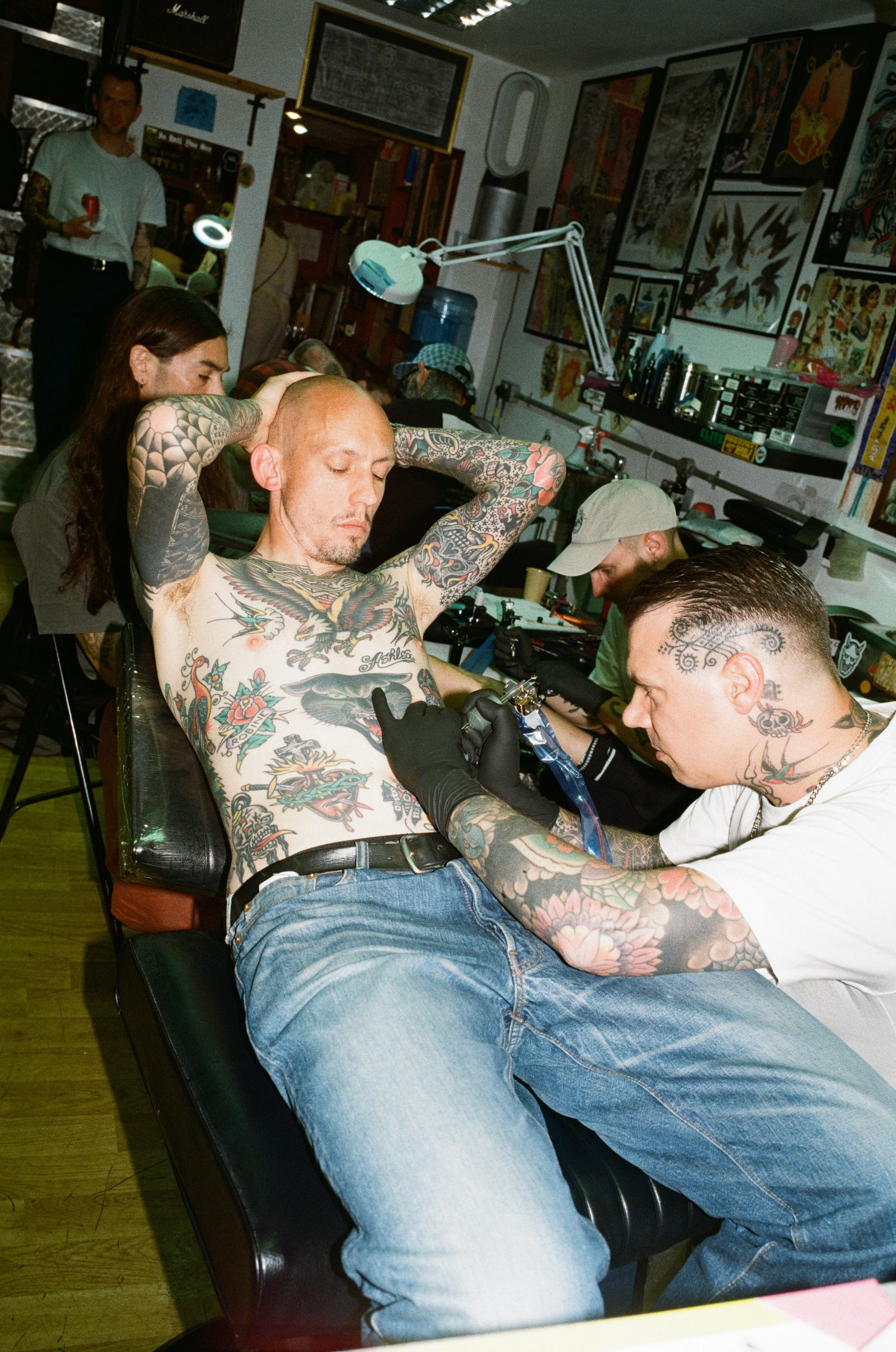 Man with multiple tattoos getting a tattoo on his chest in a tattoo shop, surrounded by artwork and other people.