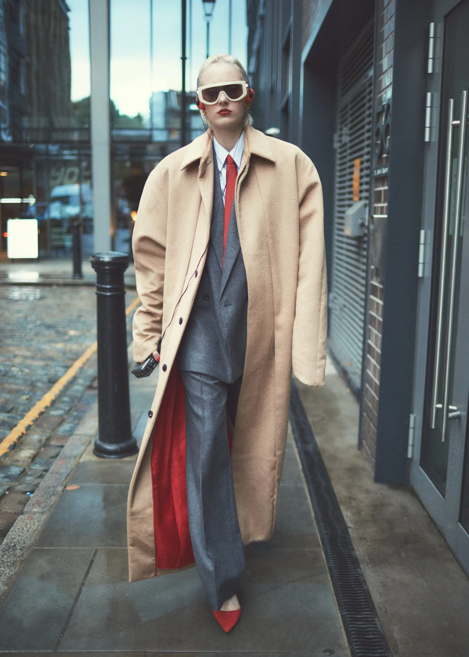 A woman walking on a city sidewalk dressed in a beige overcoat, gray pants, a gray blazer, a white shirt, and red shoes, wearing large sunglasses and listening to headphones.