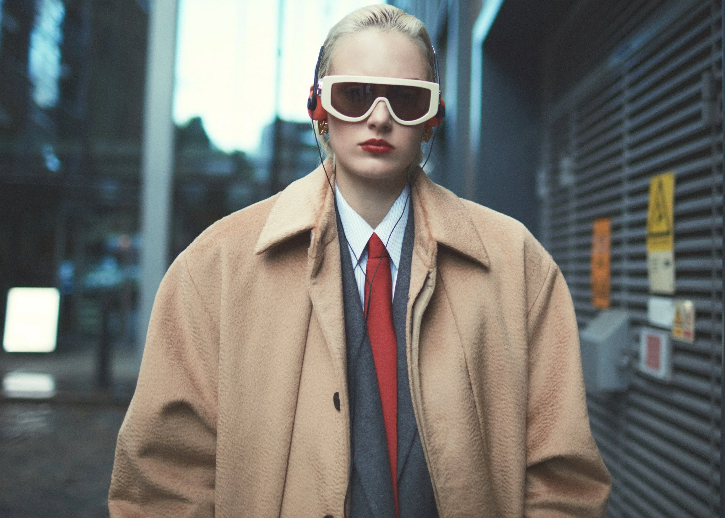 A woman with blonde hair wearing sunglasses, headphones, a beige coat, a white shirt, a red tie, and a gray vest walking in an urban environment.