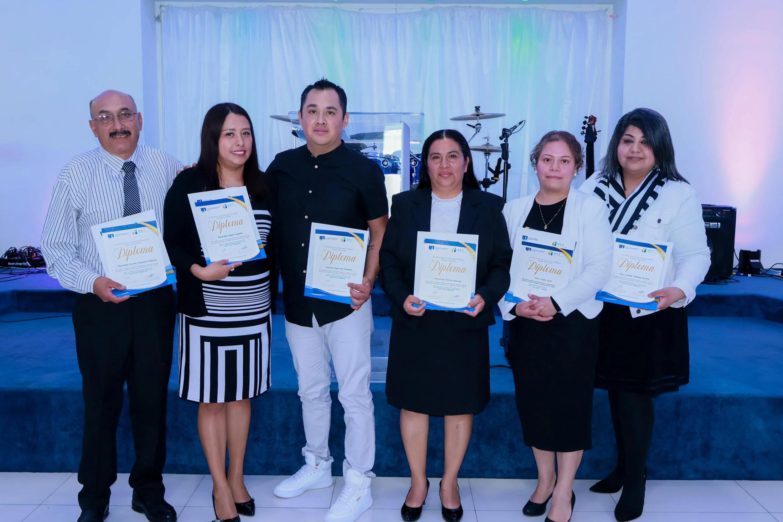 Grupo de seis personas en un escenario, sosteniendo diplomas, en una ceremonia de reconocimiento. Hay instrumentos musicales en el fondo.
