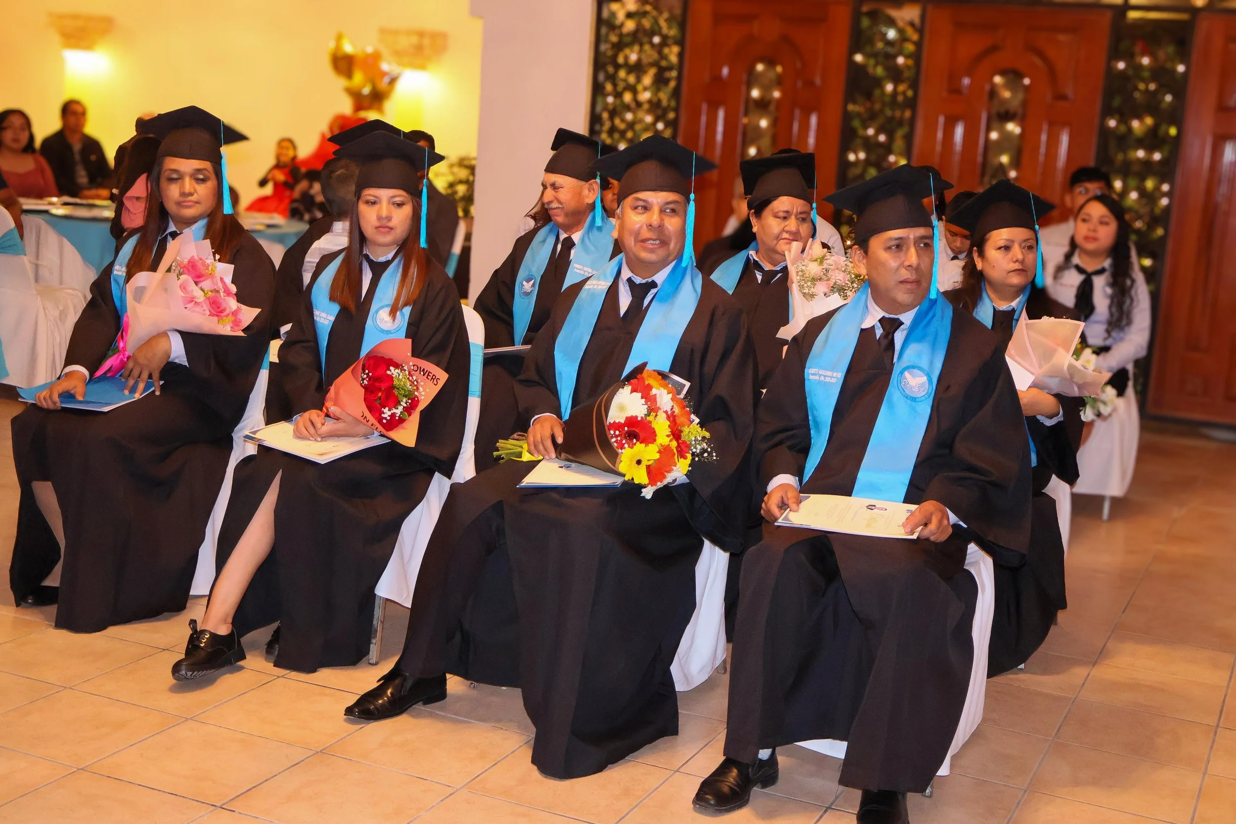 Grupo de graduados con toga y birrete en ceremonia de graduación, algunos sosteniendo flores y certificados.