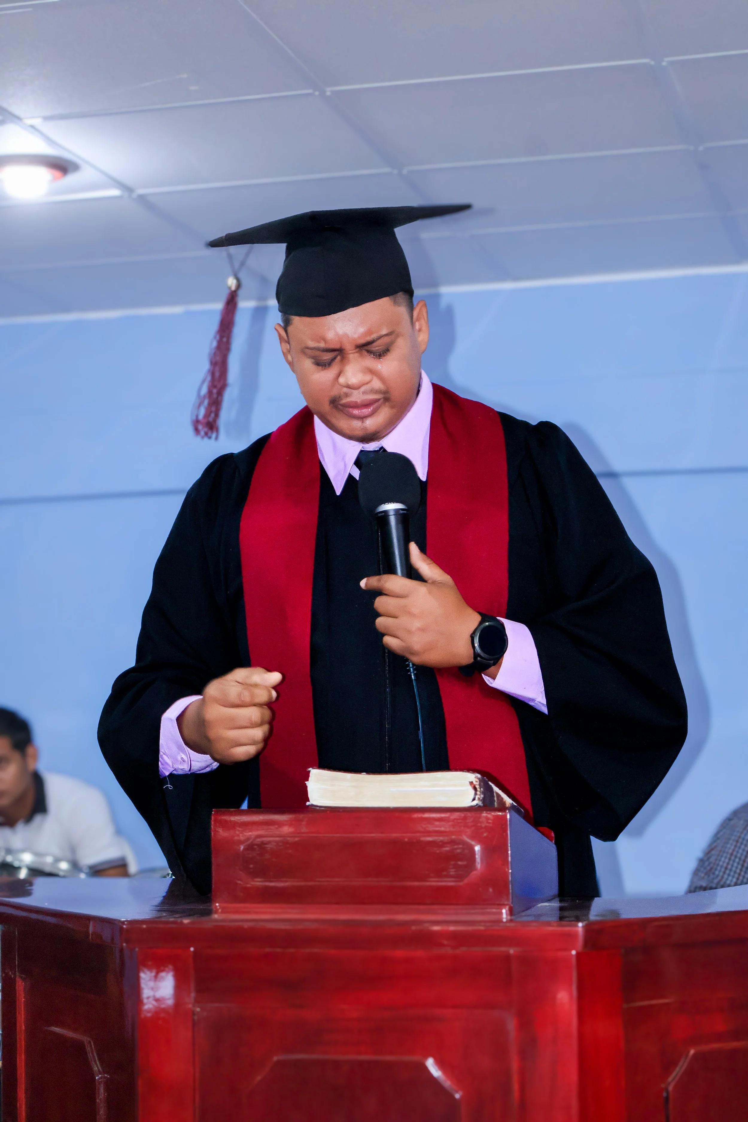 Joven vestido con toga académica y birrete en una ceremonia, con la cabeza inclinada y las manos juntas. Frente a él hay un libro en un atril de madera, mientras que en el fondo hay otras personas sentadas, y el lugar tiene paredes azules.