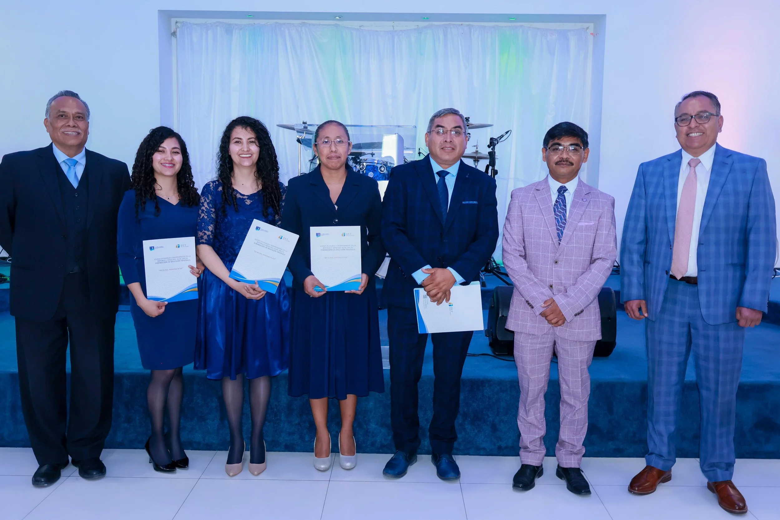 Grupo de siete personas en ceremonia formal, algunos sosteniendo certificados, en interior con piano y batería al fondo.