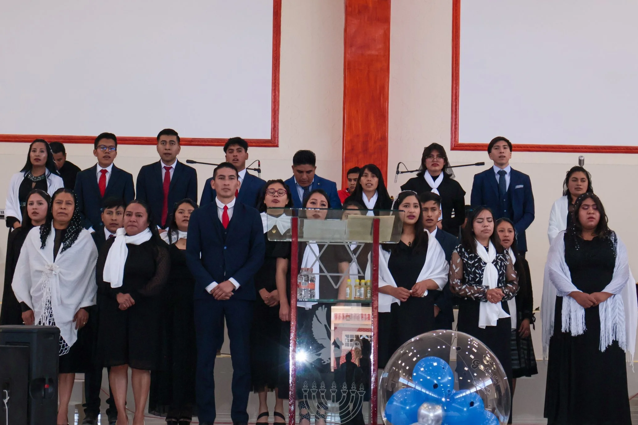 Grupo de personas en una ceremonia o evento religioso en un templo, con fondo de paredes blancas y rojas, algunos vestidos formalmente, algunos con vestimenta tradicional.