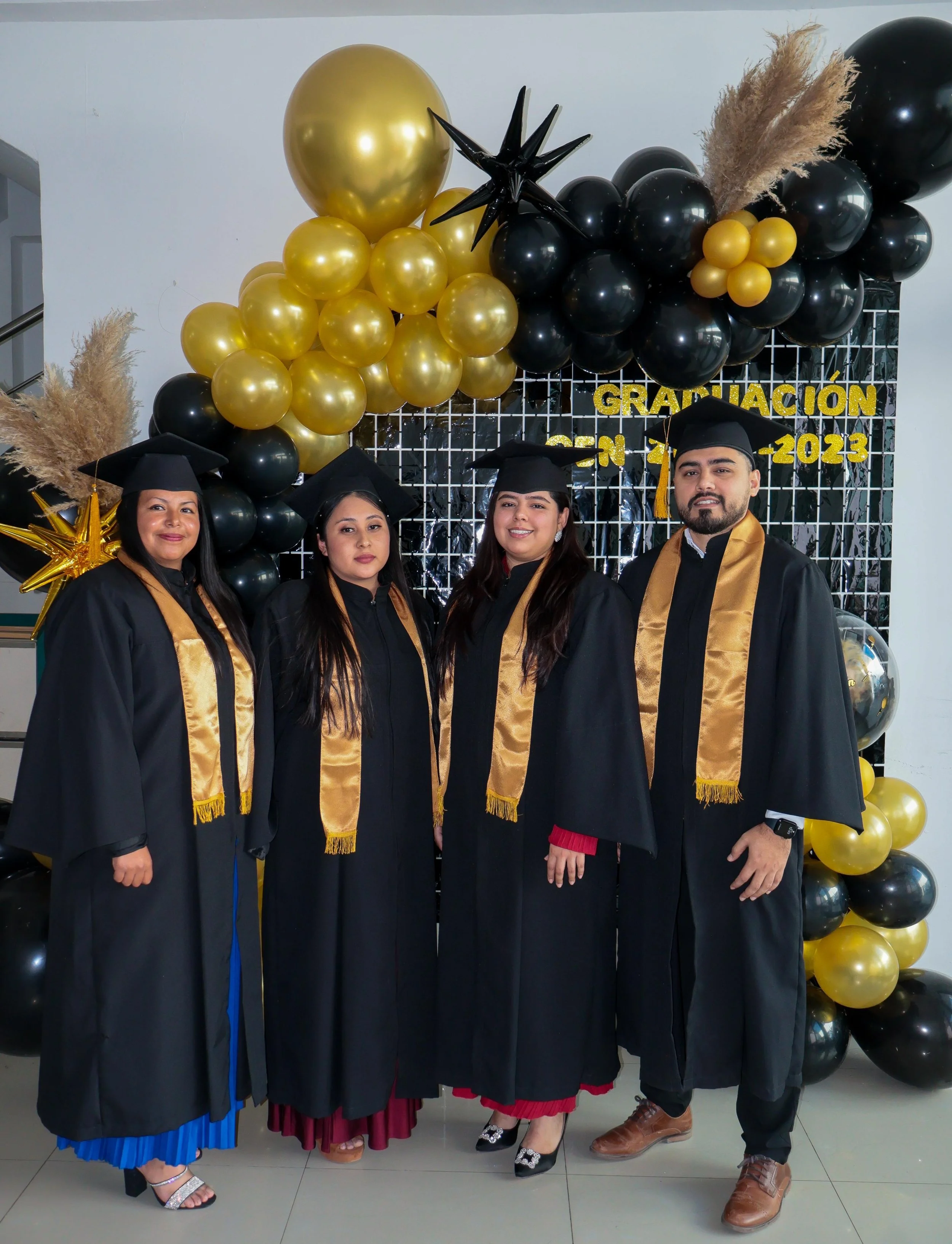 Fotografía de cuatro personas con toga y birrete de graduación en un evento de graduación, con decoración de globos en colores negro y dorado y un cartel que dice 'Graduación en 2023'.