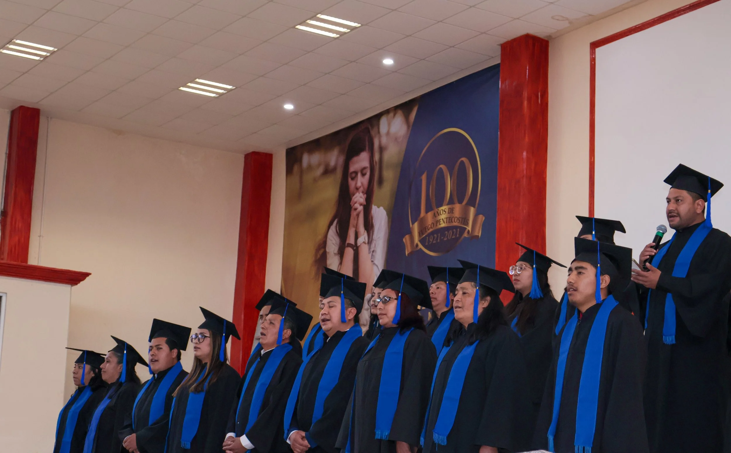 Grupo de graduados en ceremonia de graduación, usando toga y birrete, con un cartel conmemorativo en el fondo.