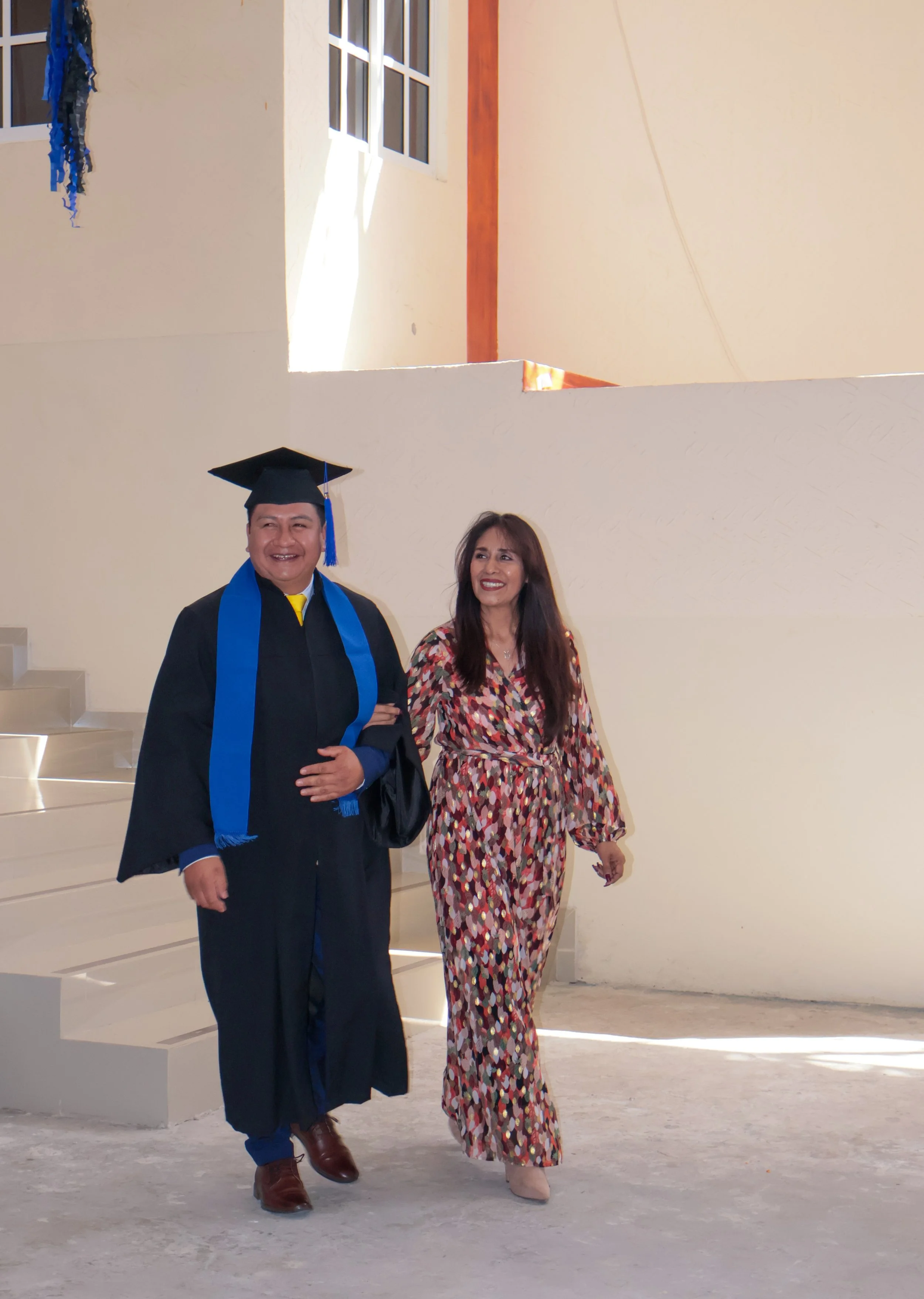 Persona en toga de graduación con otra persona en un ambiente interior, con paredes claras y ventanas, en una celebración académica.