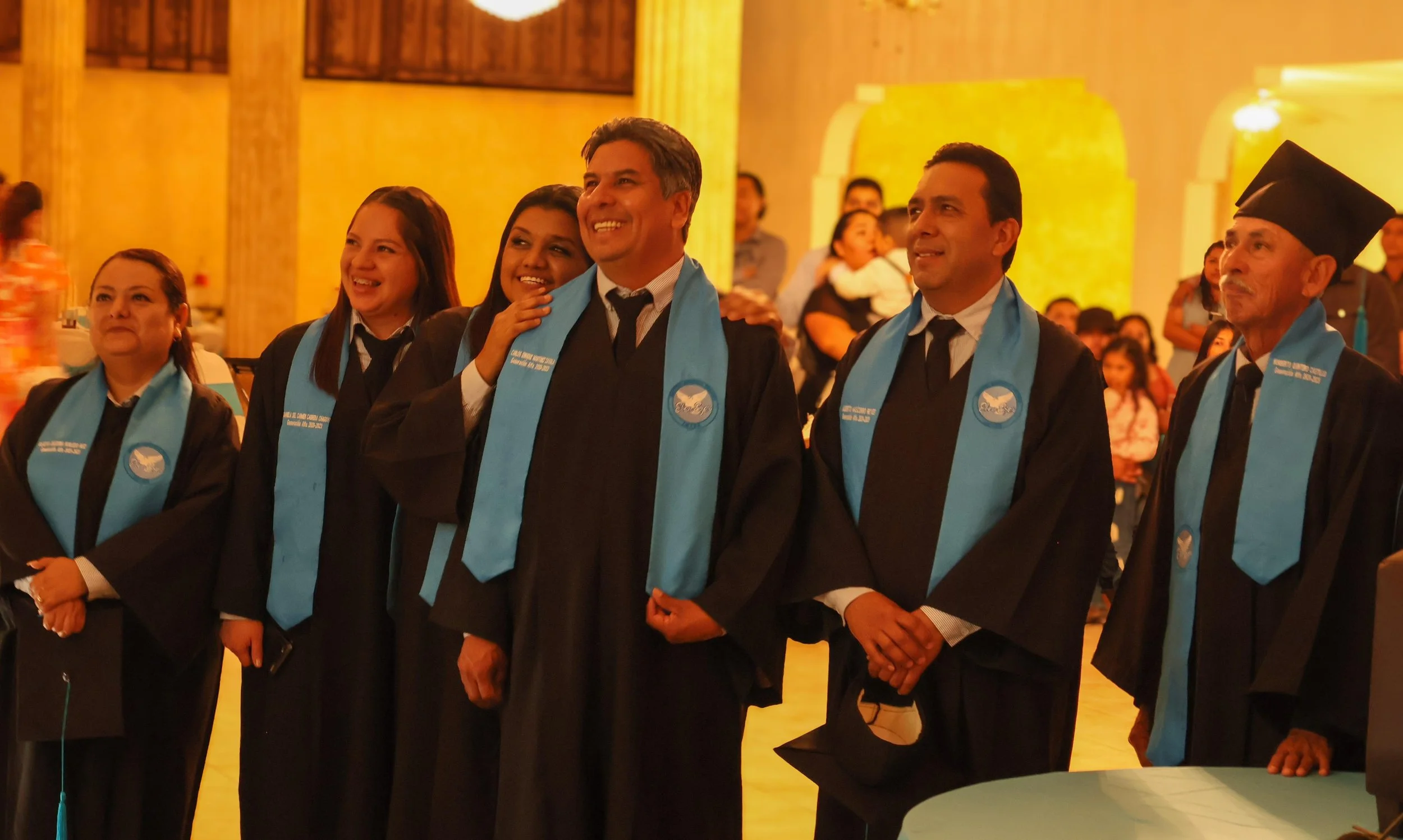 Grupo de personas en toga y birrete durante una ceremonia de graduación en un salón con fondo dorado y público