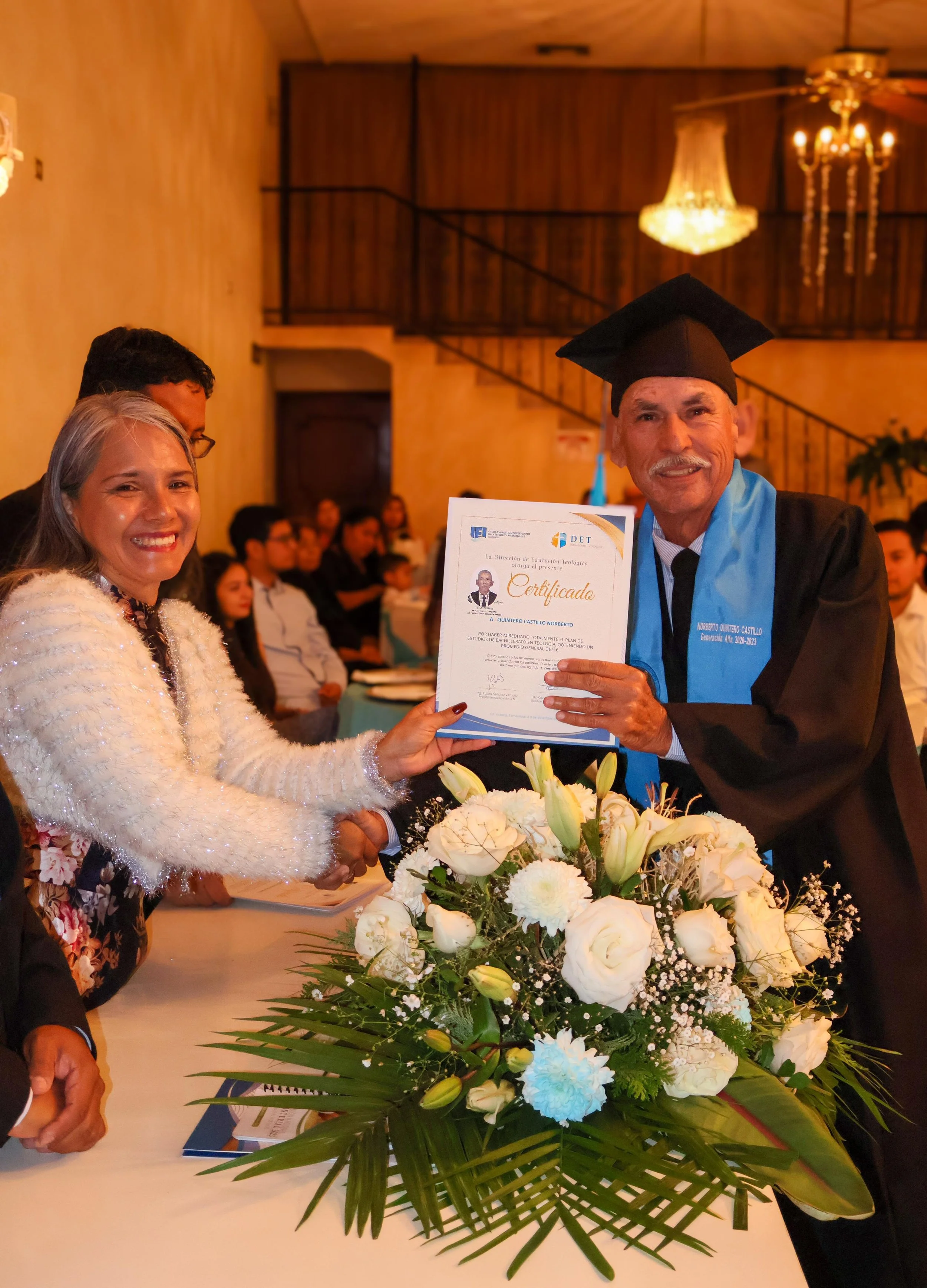 Persona en ceremonia de graduación sosteniendo un certificado, acompañada de otros asistentes, con un arreglo floral en primer plano, en un salón decorado con elegancia.