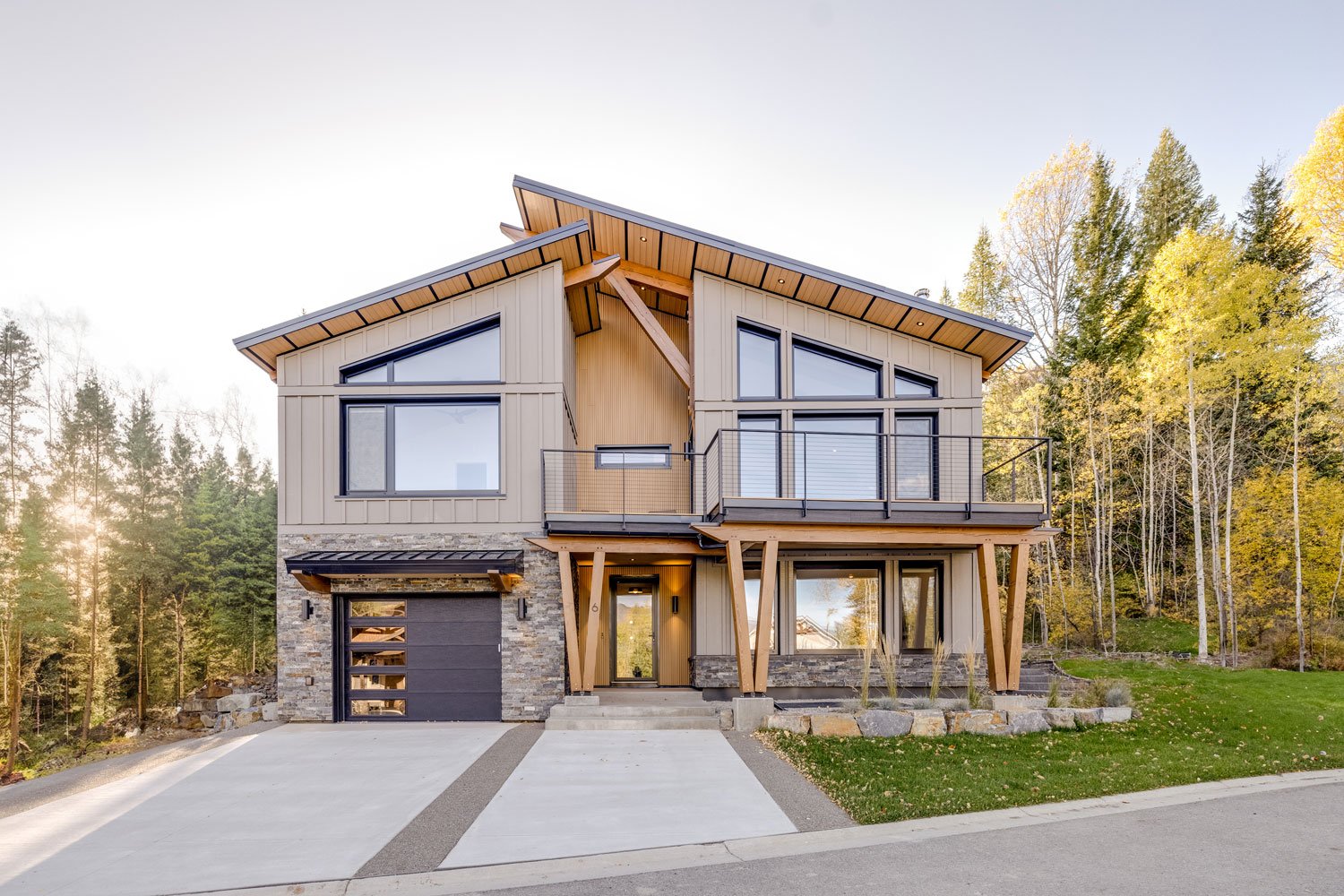 Modern two-story house with a mix of wood, stone, and metal exterior, large windows, a small front porch, and a garage, set against a backdrop of trees and a clear sky in daylight.