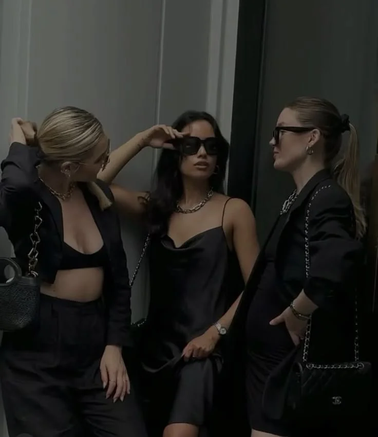 Tres mujeres jóvenes posando juntas en un entorno urbano o de moda, vestidas con elegantes prendas negras y accesorios de joyería.
