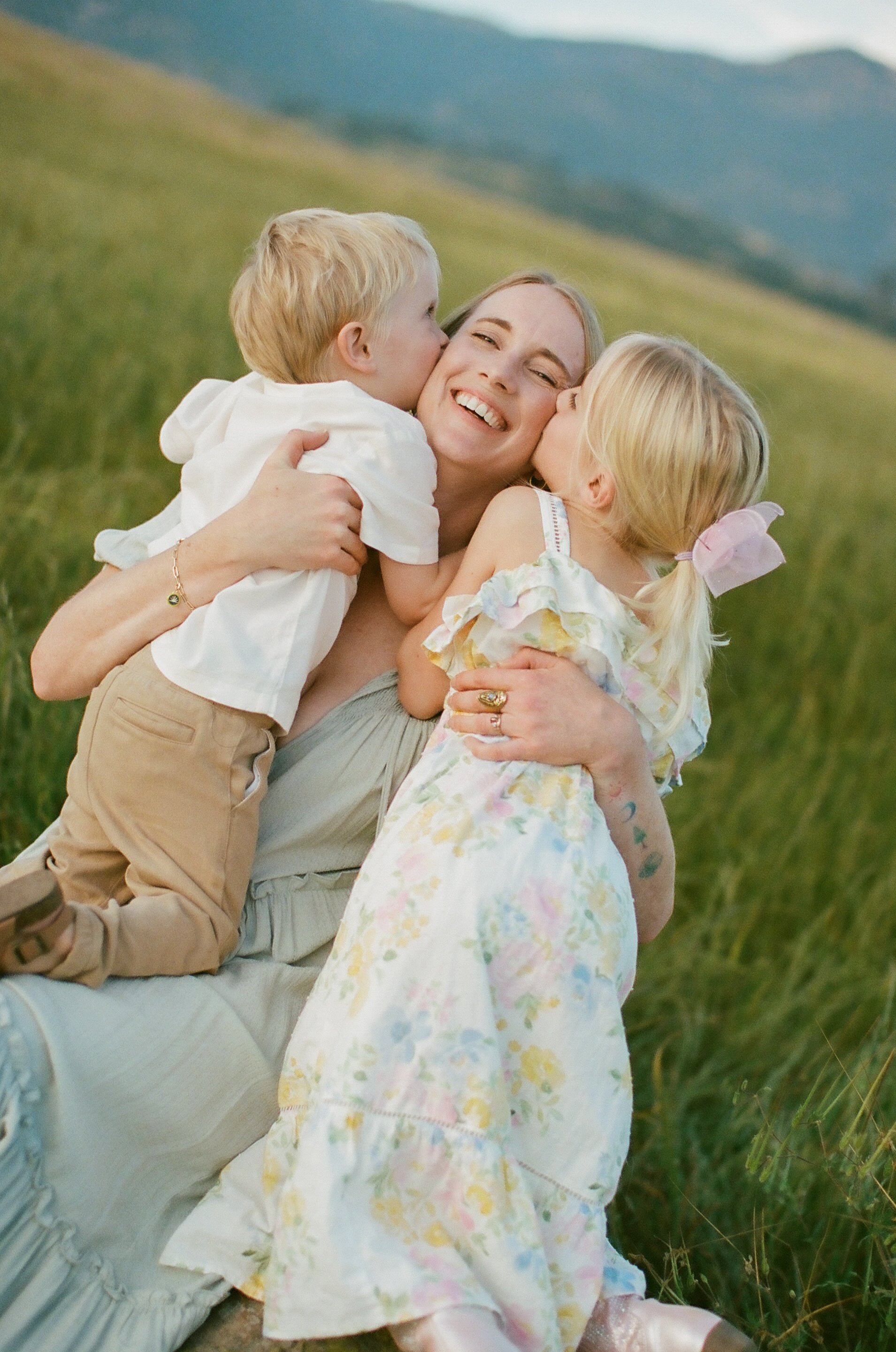 A woman smiling while hugging two children, a boy and a girl, outdoors in a grassy area during daytime.