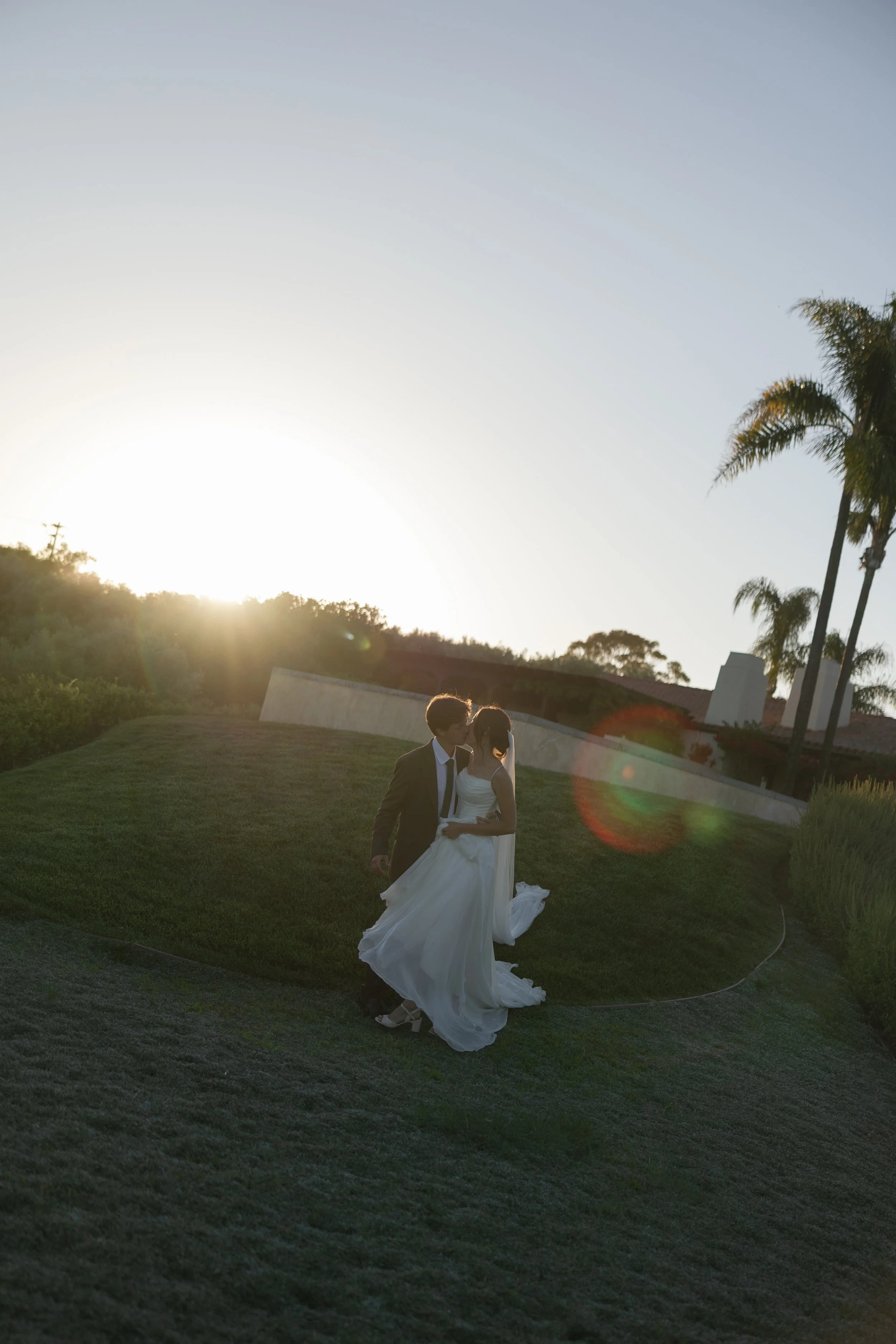 A couple dressed in wedding attire standing on a grassy hill during sunset, with palm trees in the background and the sun casting a glow.