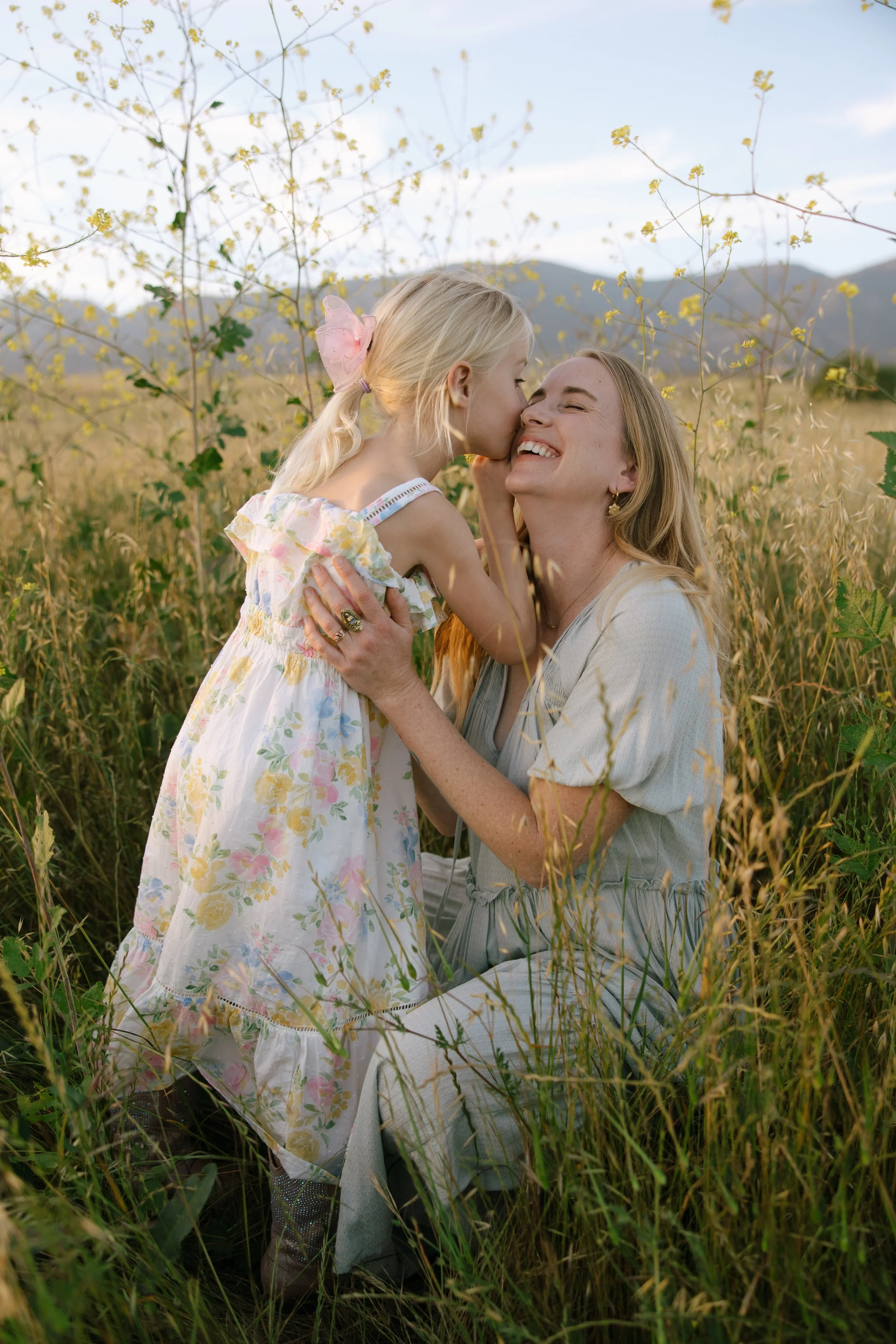 A woman and a young girl sharing a moment of affection in a wheat field, with mountains and a blue sky in the background.