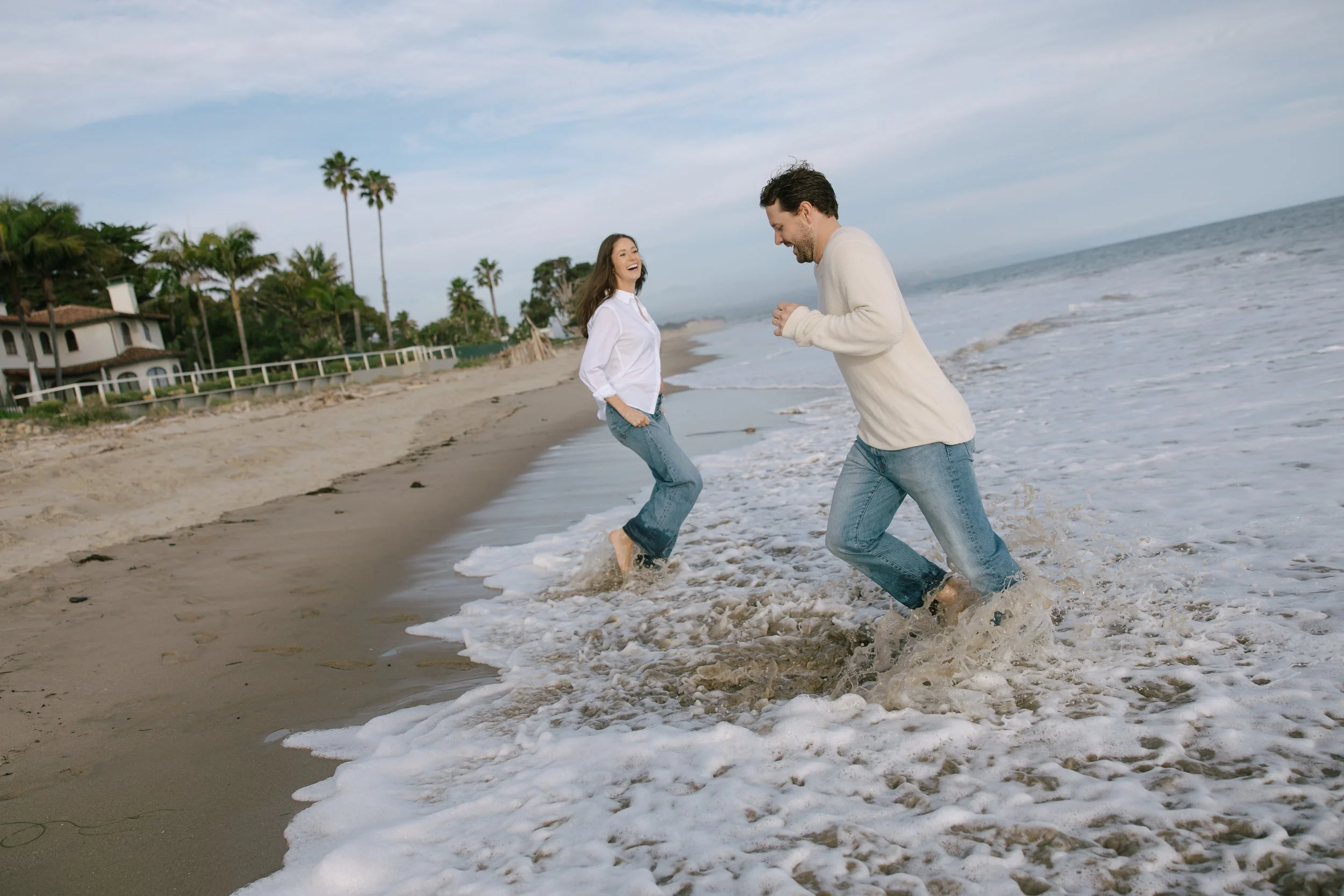 A man and woman laughing and playing in the ocean waves at the beach, with palm trees and houses in the background under a partly cloudy sky.