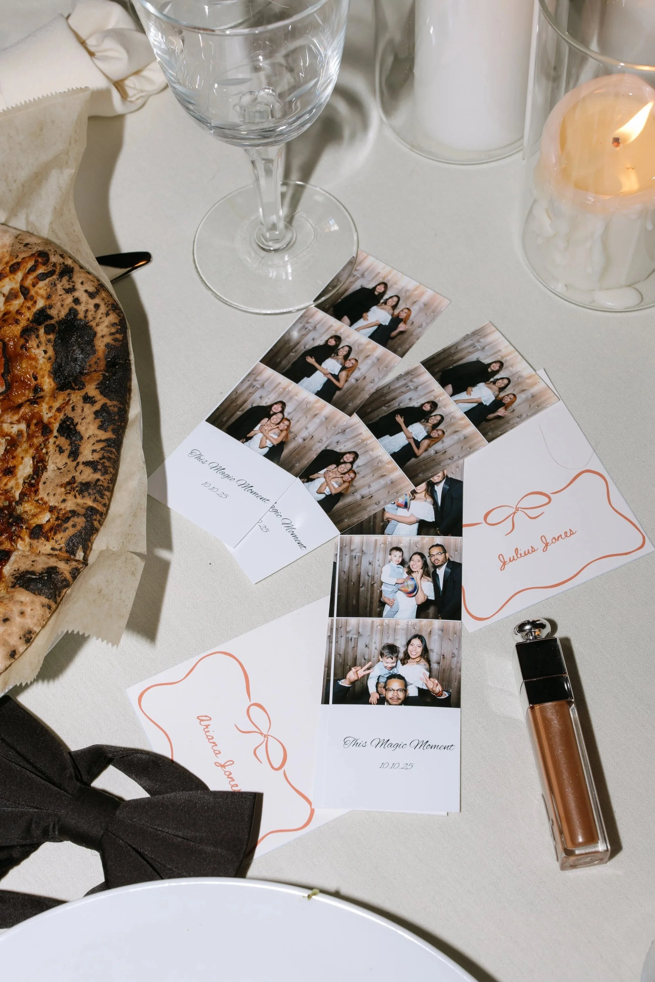 A table setting with a pizza, empty wine glass, decorative candles, several photo booth strips, a wedding invitation, and a lipstick tube.