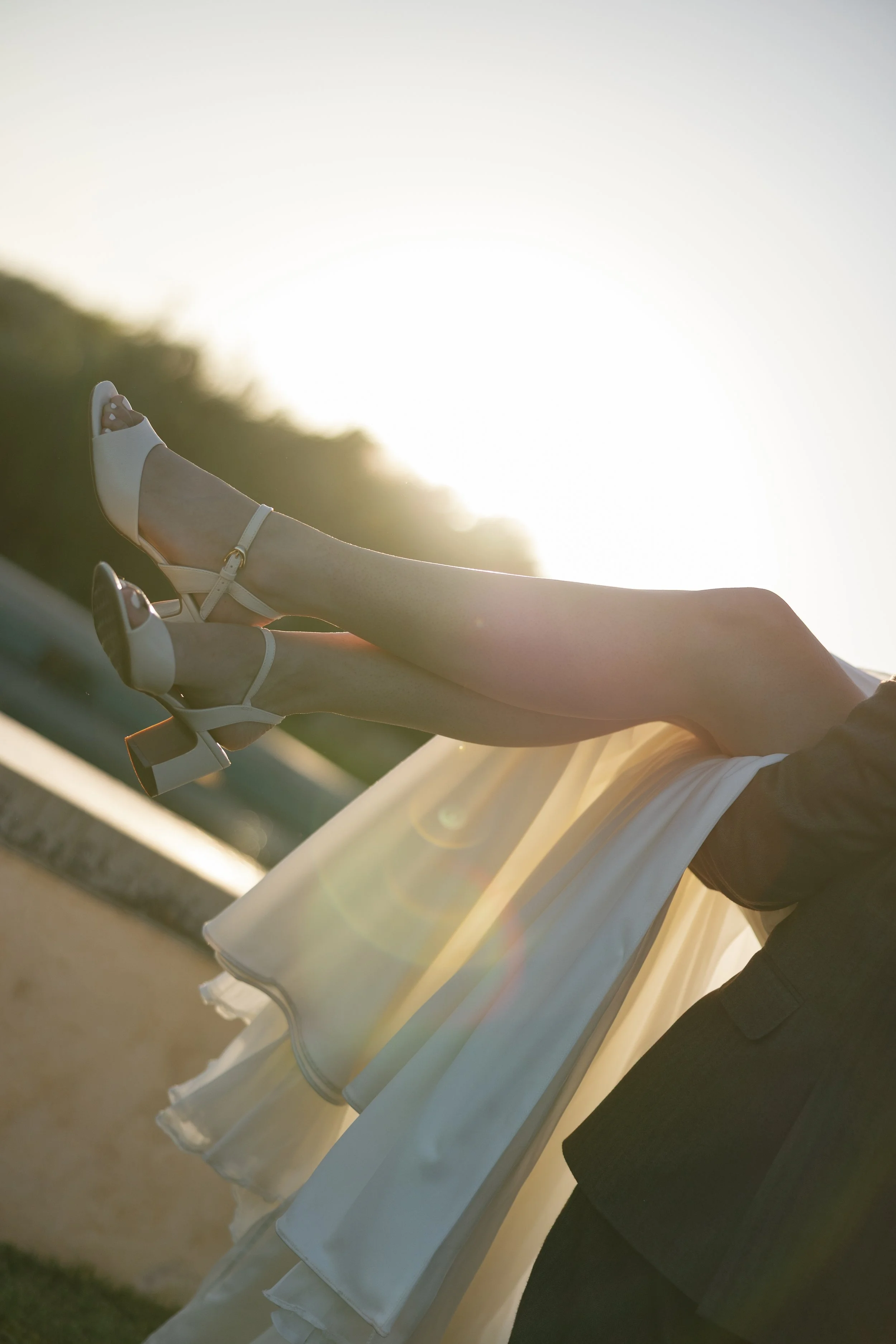 Close-up of a person's legs and feet in white high-heeled shoes, with a flowing white dress, taken outdoors with sunlight creating a warm glow.