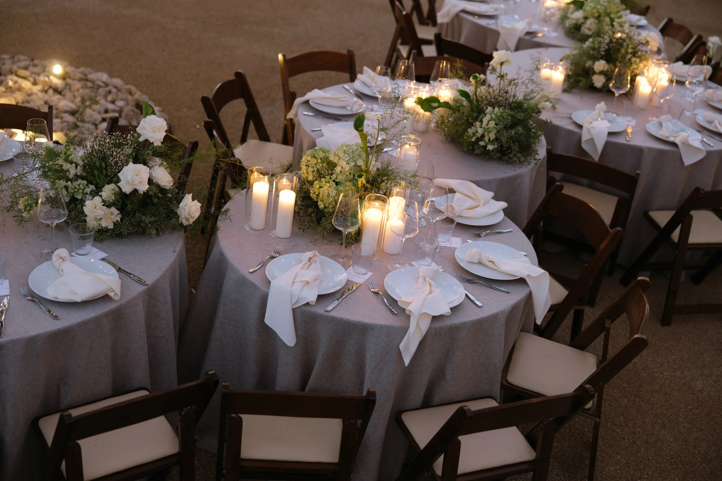 A decorated outdoor dining table with white linen tablecloths, floral centerpieces, and candlelit candles at sunset or evening, set for a formal event.