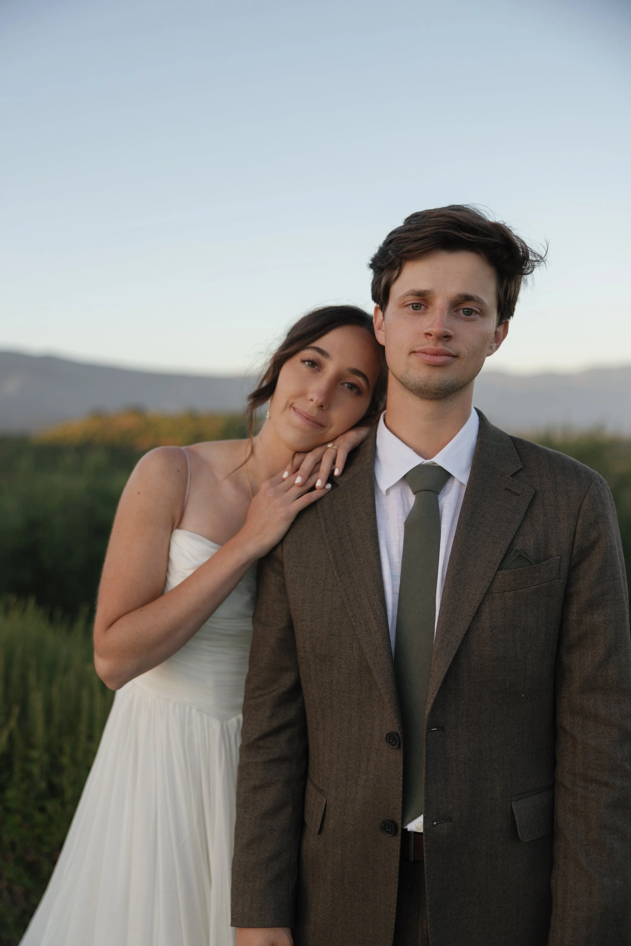A young woman with dark hair and a white dress is resting her head on the shoulder of a young man with brown hair wearing a suit and tie, outdoors in a field with mountains in the background during the daytime.