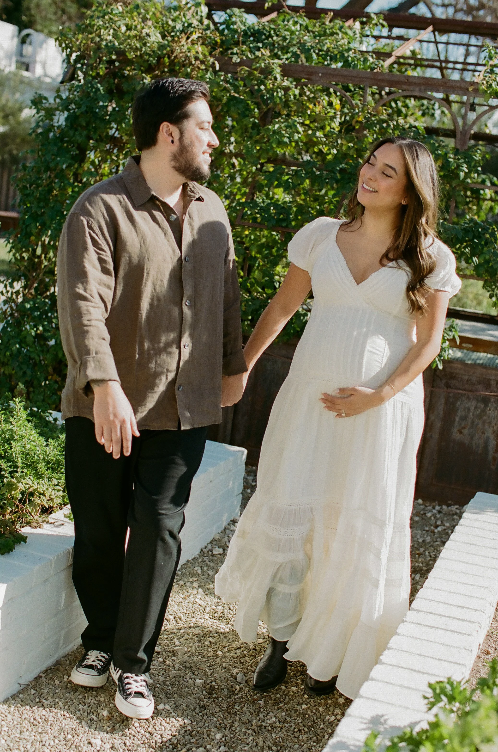 A pregnant woman in a white dress and black boots smiling and holding her belly, standing with a man in casual clothing and sneakers, outdoors near white brick planters and green bushes.