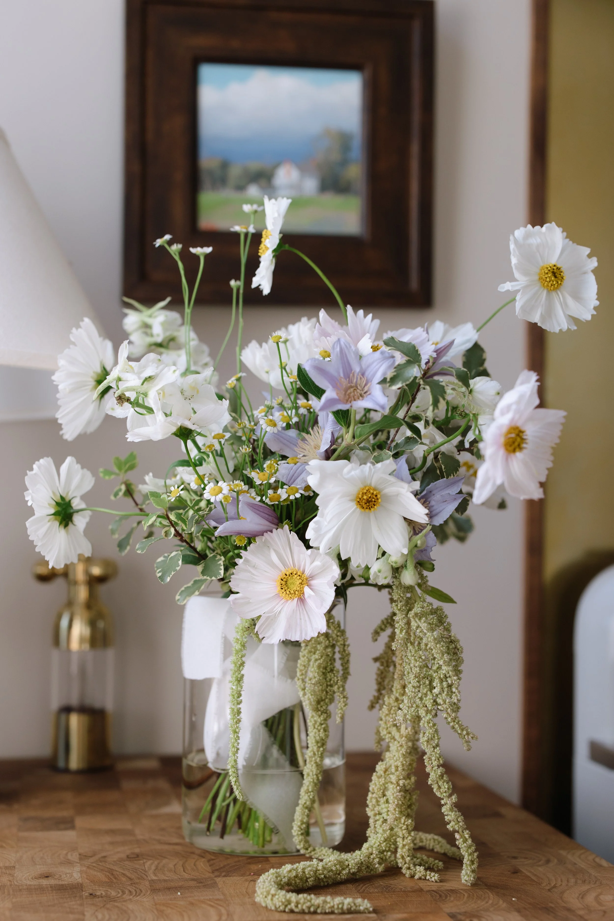 A floral arrangement with white flowers and long hanging greenery in a glass vase on a wooden table, with a framed landscape painting in the background.