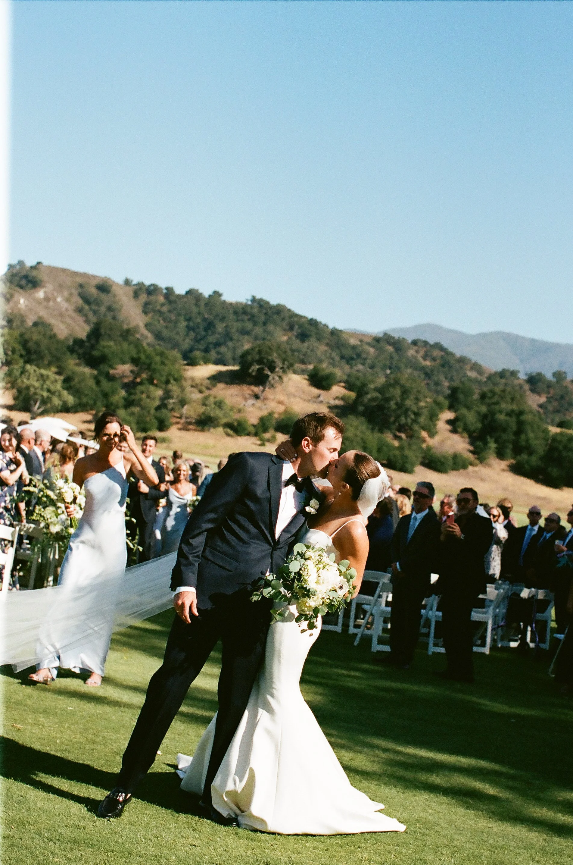 A bride and groom sharing a kiss during an outdoor wedding ceremony, with guests seated and standing behind them, set against a backdrop of rolling hills and clear blue sky.
