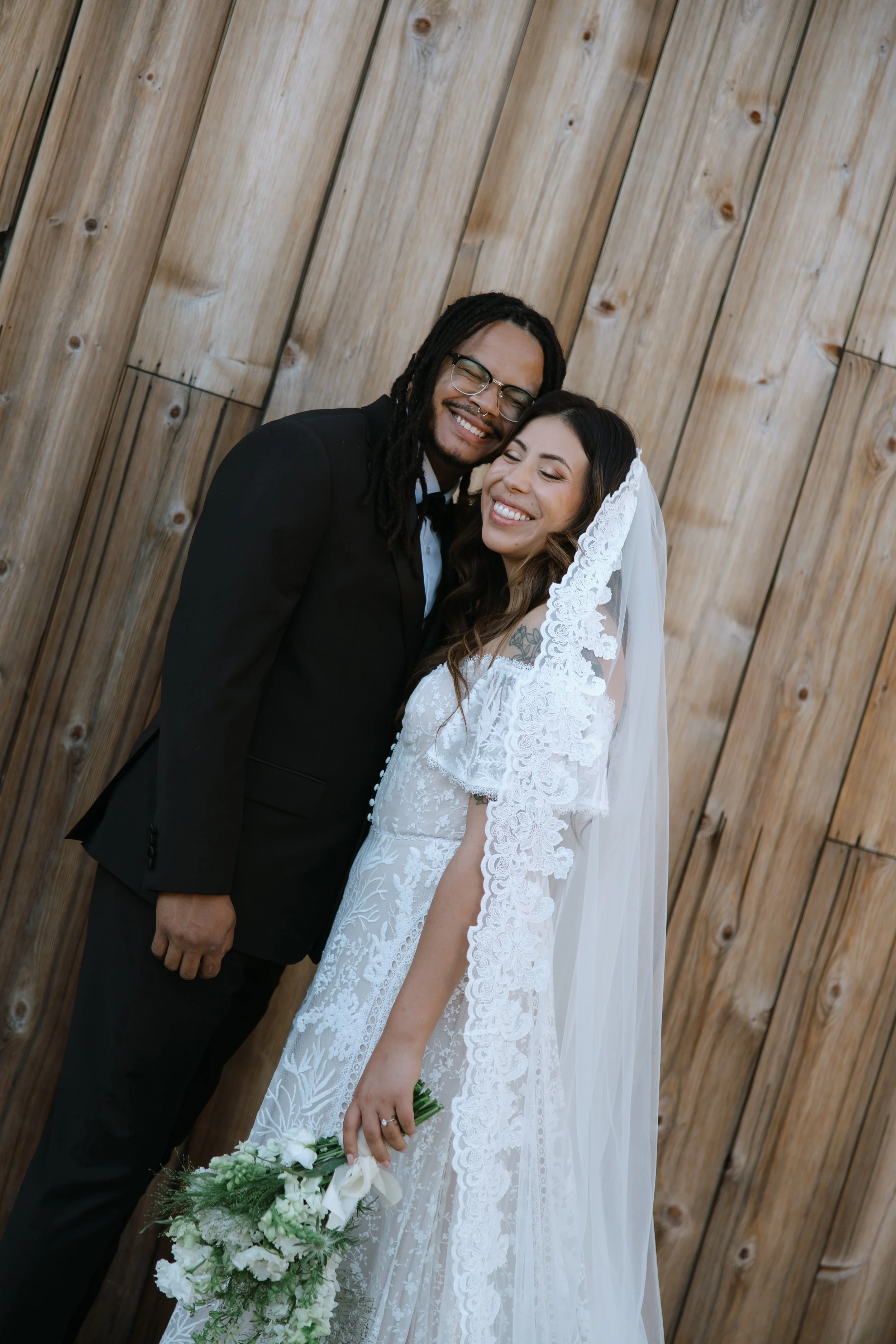 A smiling couple, the groom with dreadlocks and glasses in a tuxedo, and the bride with long brown hair and a lace wedding gown, standing close together and holding a bouquet of white flowers, in front of a wooden wall.