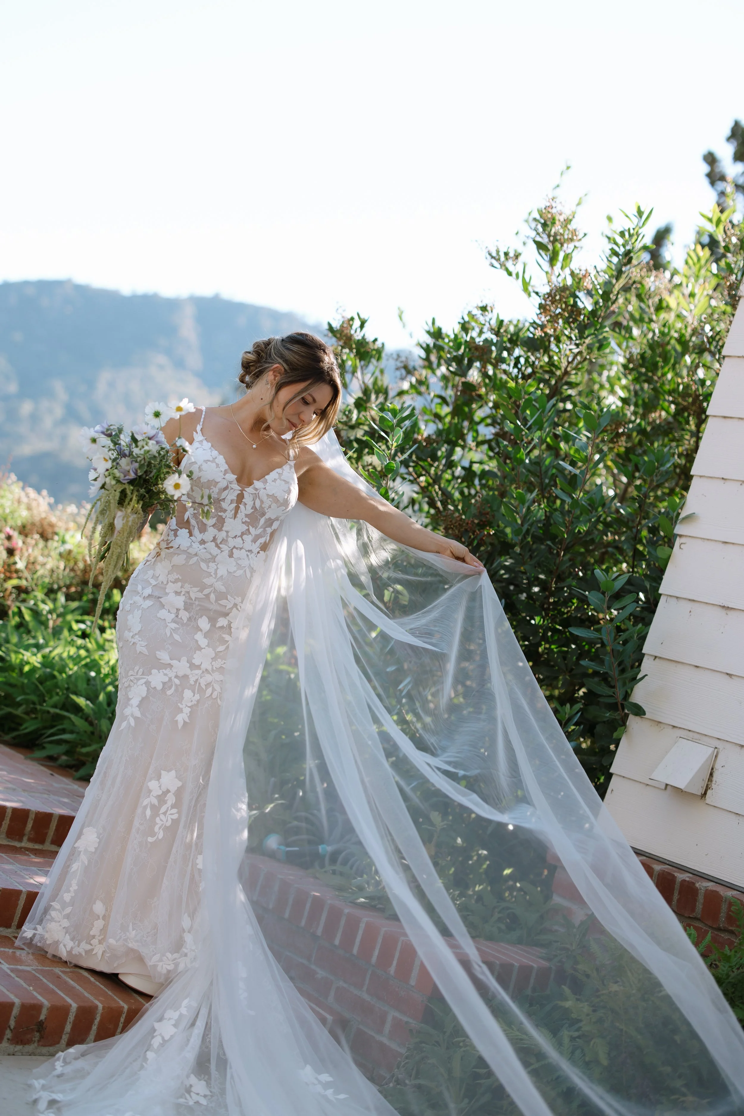 Bride in a lace wedding gown holding a bouquet, outside on brick steps with greenery and a mountain backdrop.